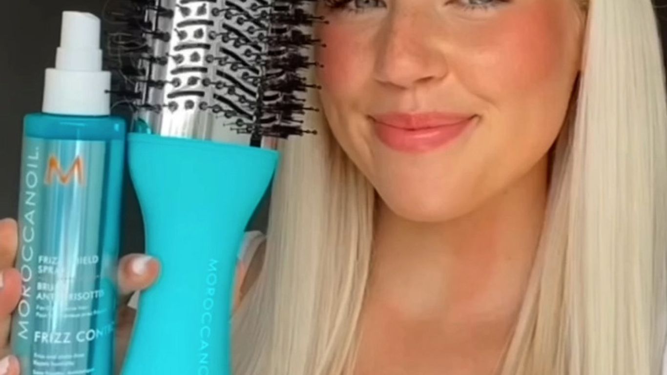 Woman smiling, holding hairbrush and hair product, beauty tutorial vibe.