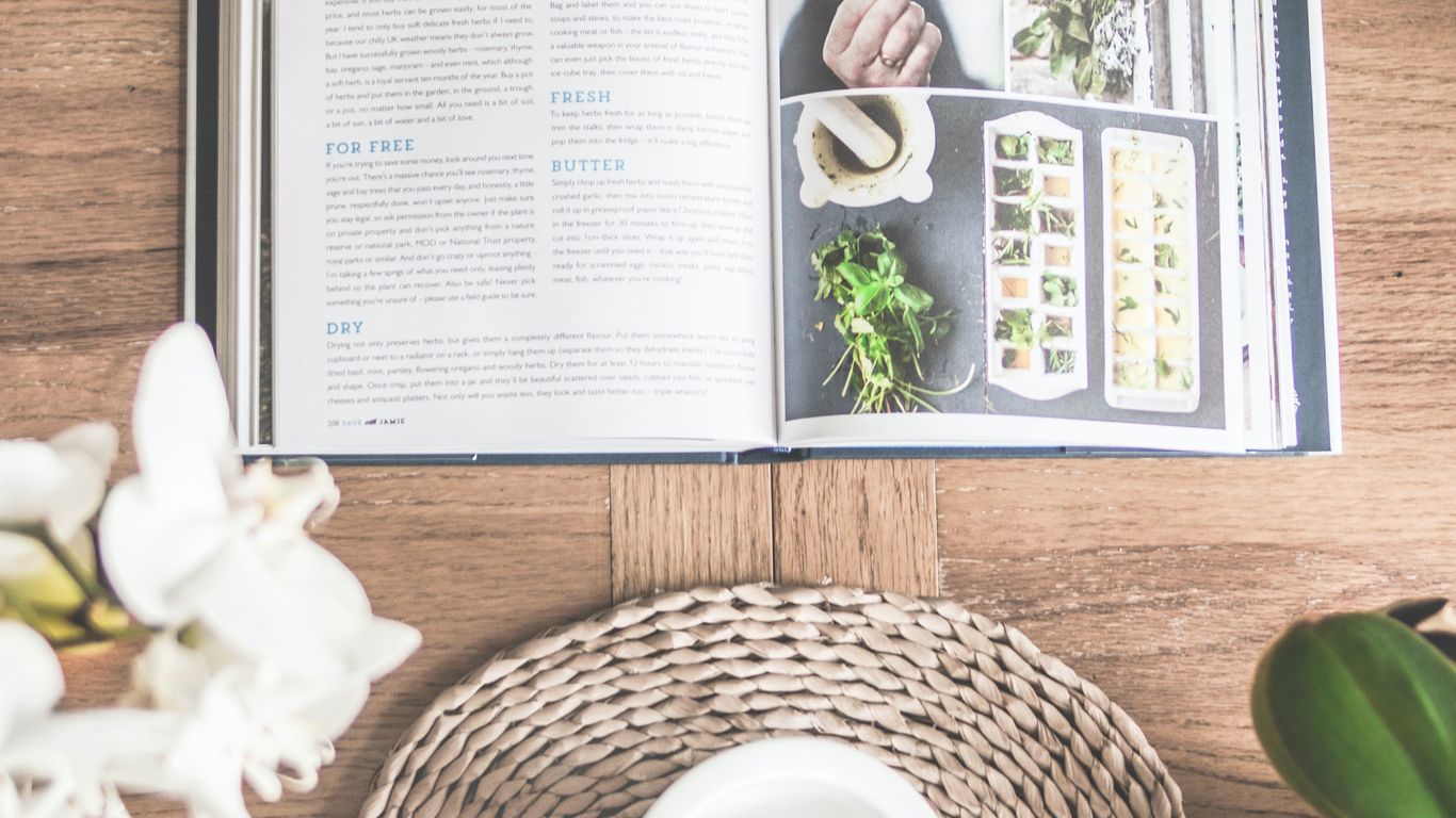 Open cookbook about herbs, mortar and pestle, wooden table.