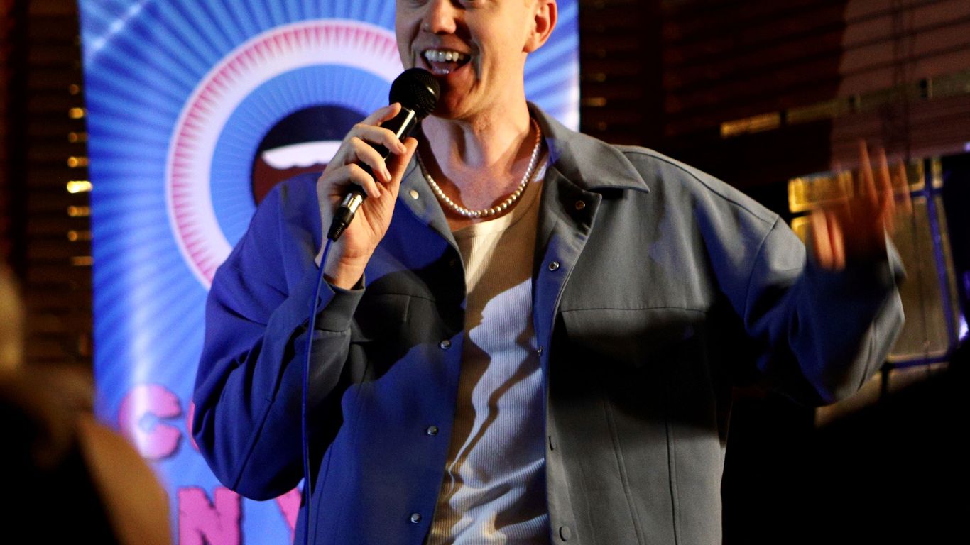 A person holds a microphone and speaks on stage with a banner.