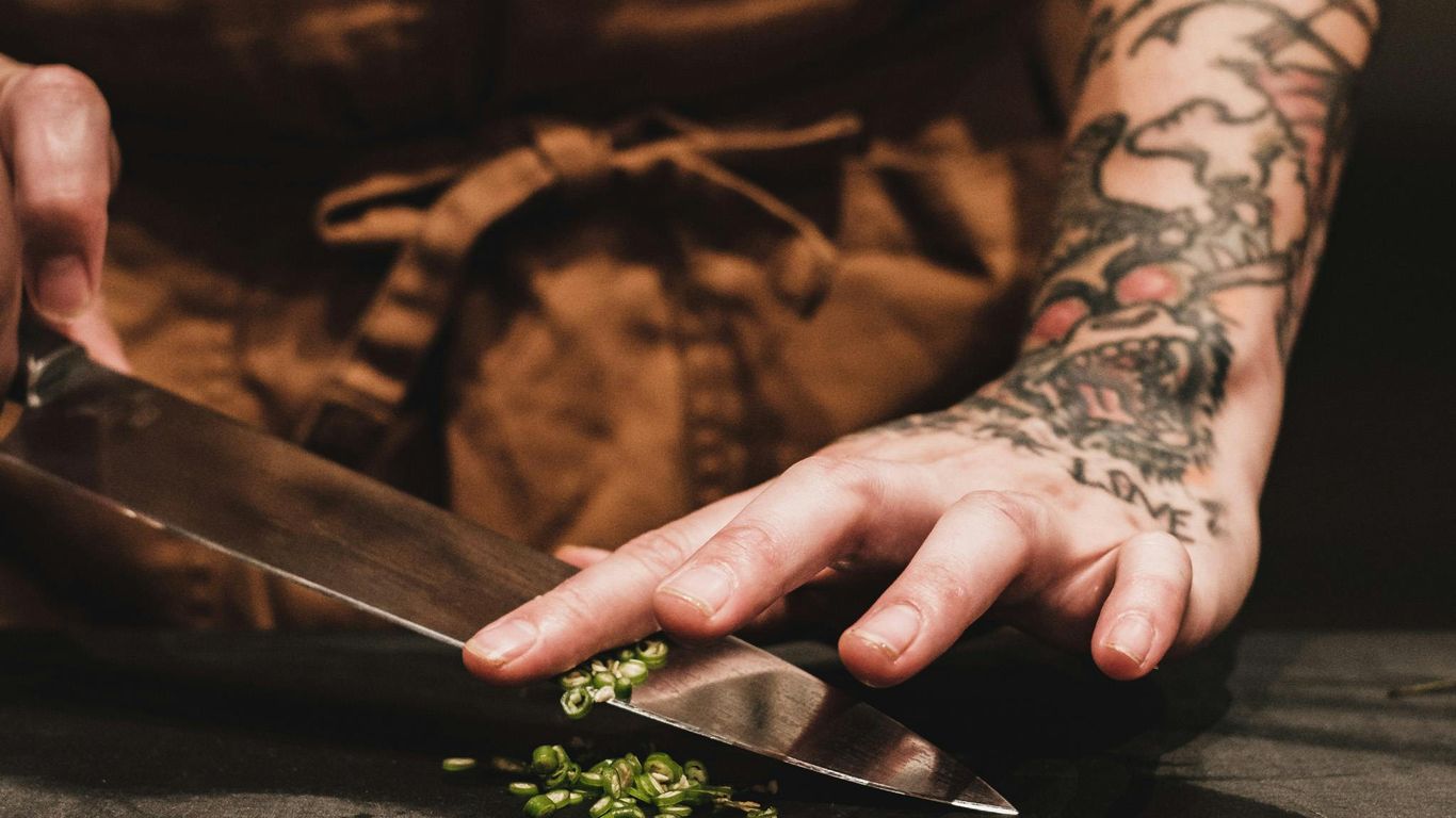 Tattooed person chopping green herbs with a large knife.
