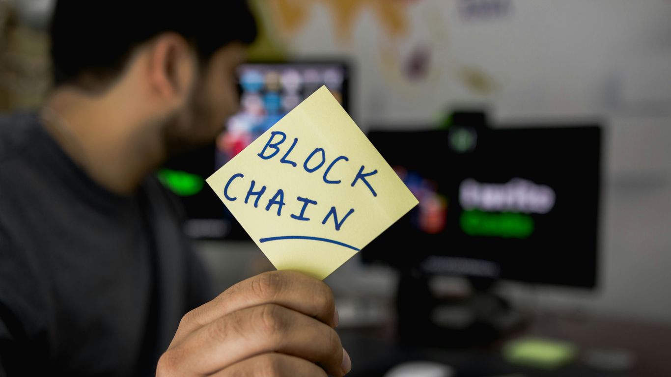 Person holding a sticky note labeled "BLOCKCHAIN" in an office.