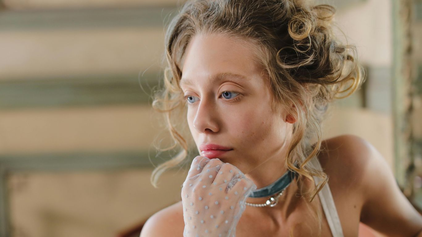 A woman with blue eyes and blonde hair wears a choker and gloves.