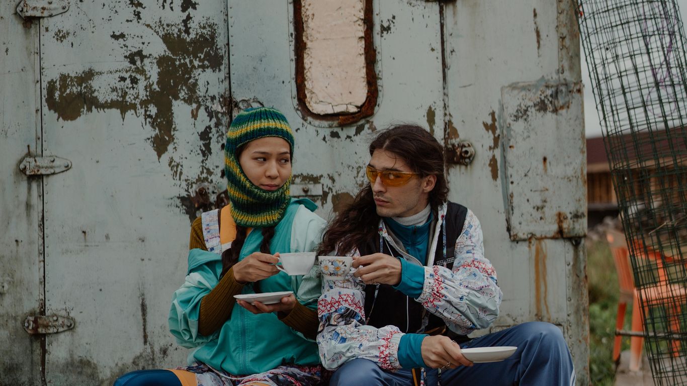 Two people in colorful outfits sit outside, drinking tea together.