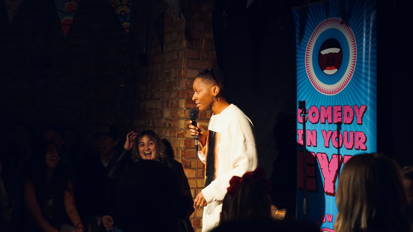 A person performs stand-up comedy on stage with a banner.