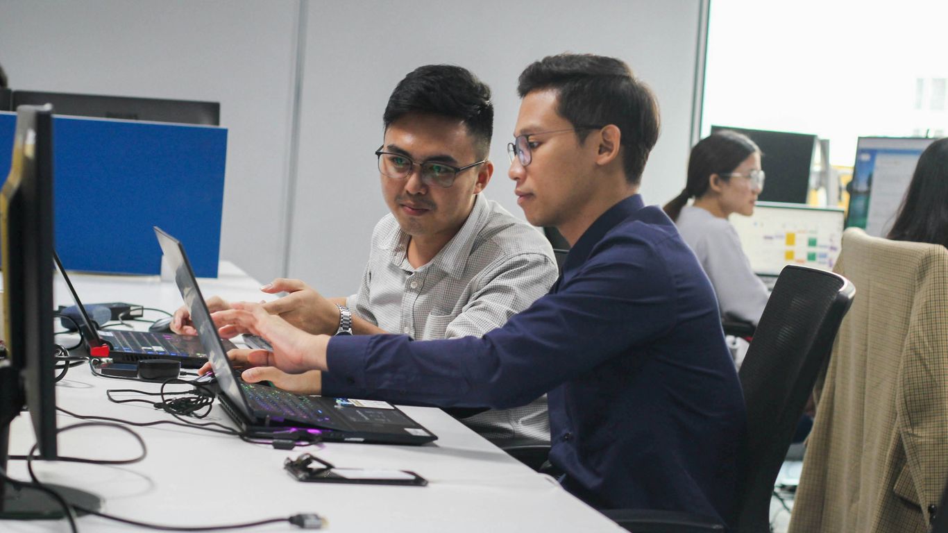 two men sitting at a desk looking at a laptop