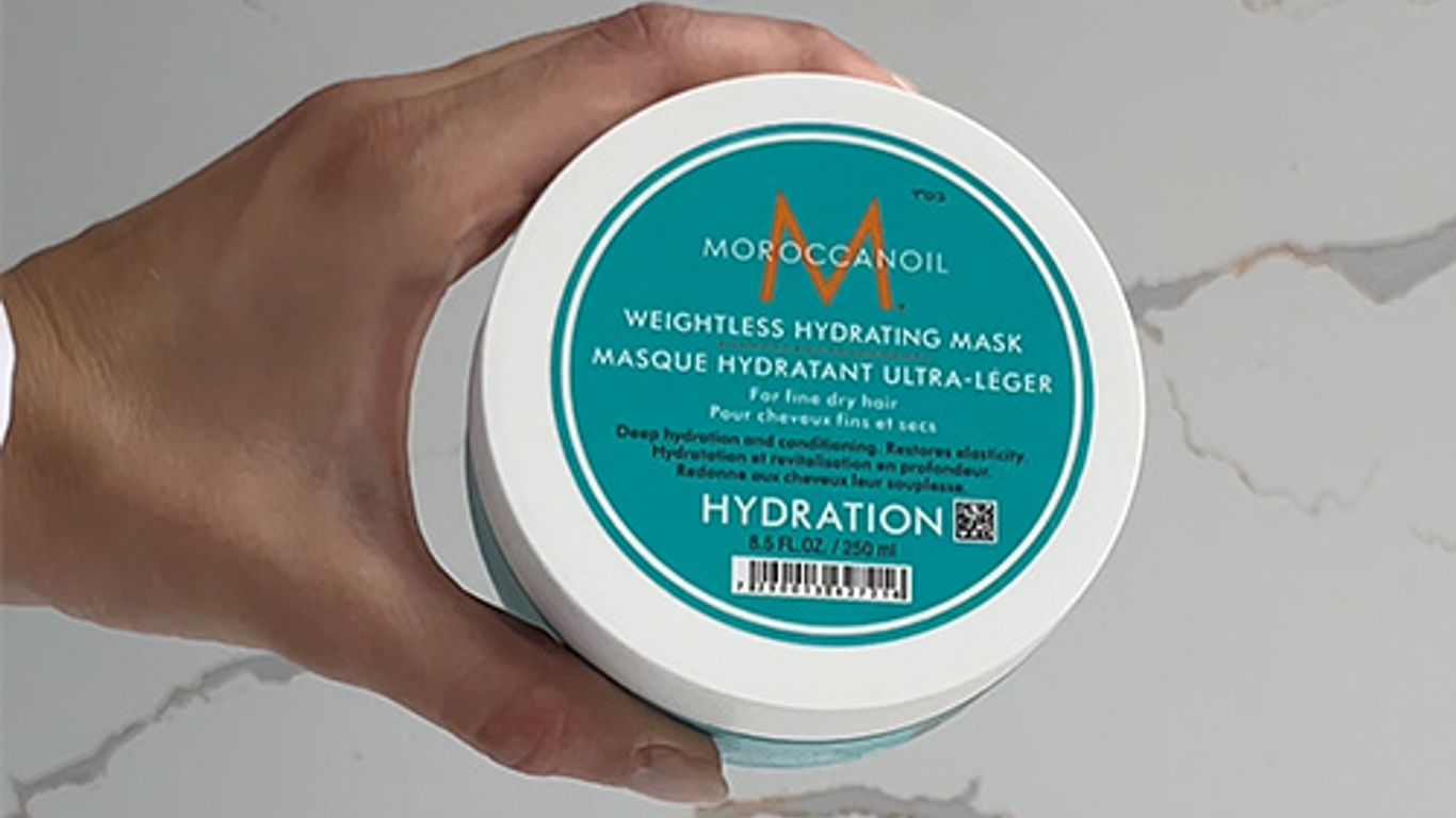 Hand holding a MoroccanOil hydrating hair mask container on marble.