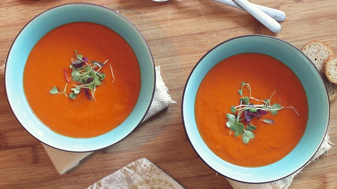 Two bowls of tomato soup with garnish, bread, and spoons.