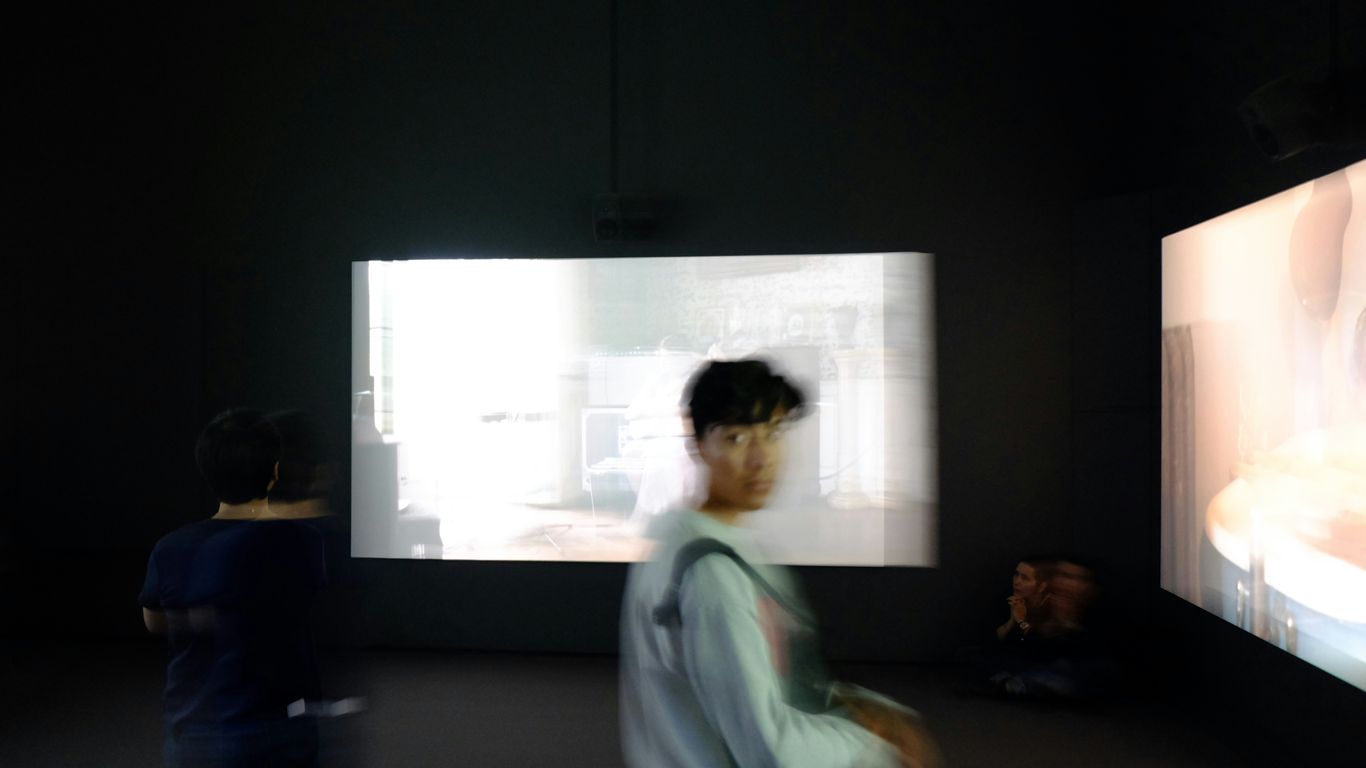 person in grey sweater standing in front large monitors