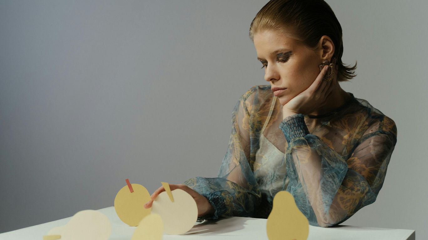Person with hand on chin near paper fruit shapes on table.