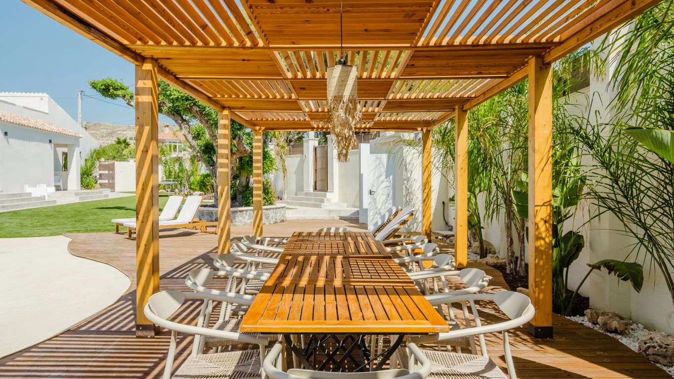 Wooden pergola with dining table and chairs outdoors.