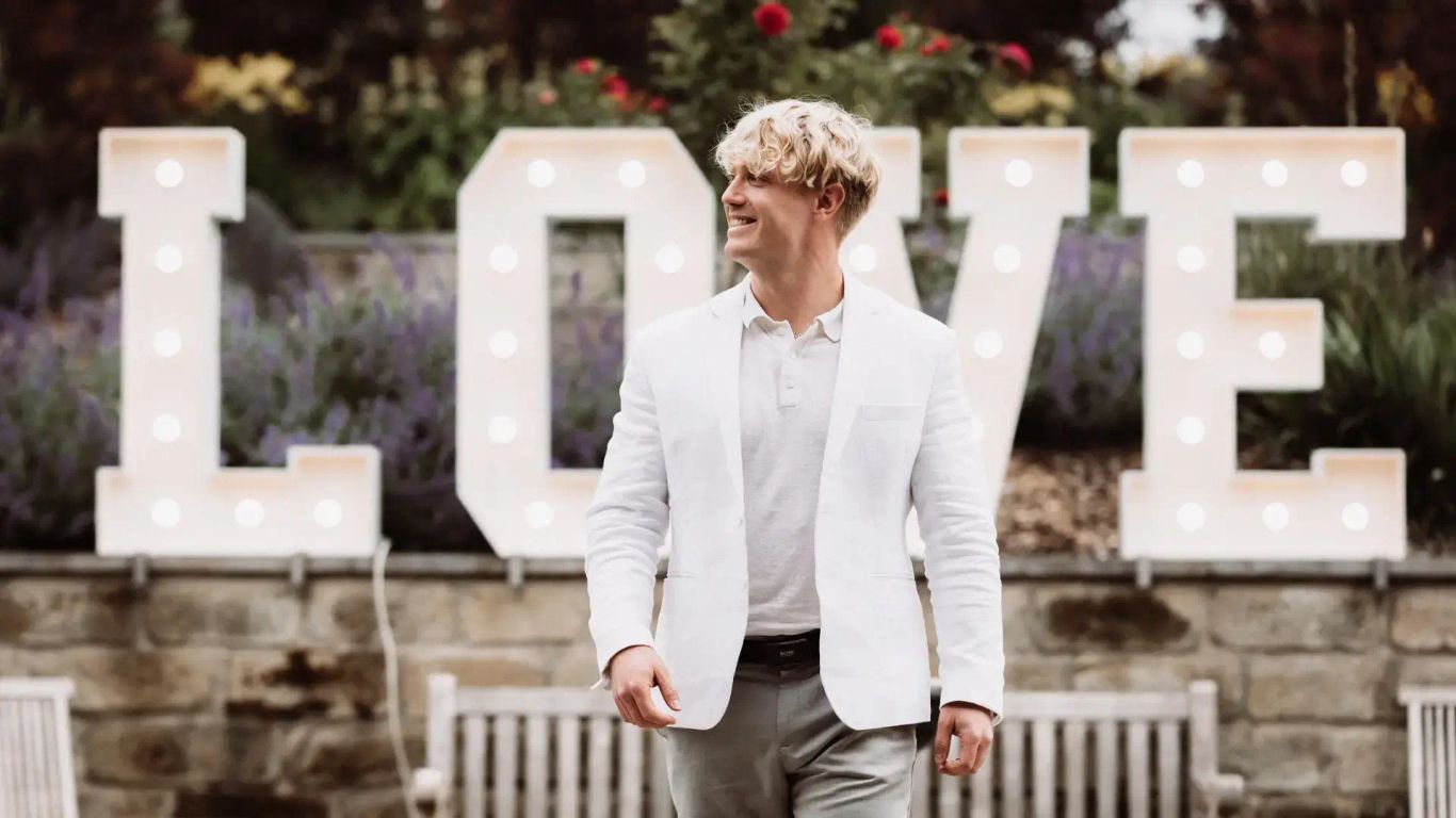Nathan White im weißen Anzug, lächelnd, mit einem großen ‚LOVE‘-Schriftzug im Hintergrund.