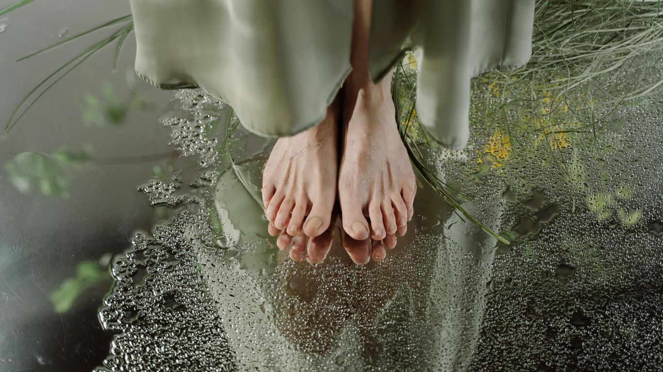 Bare feet on wet surface with reflection, surrounded by greenery.