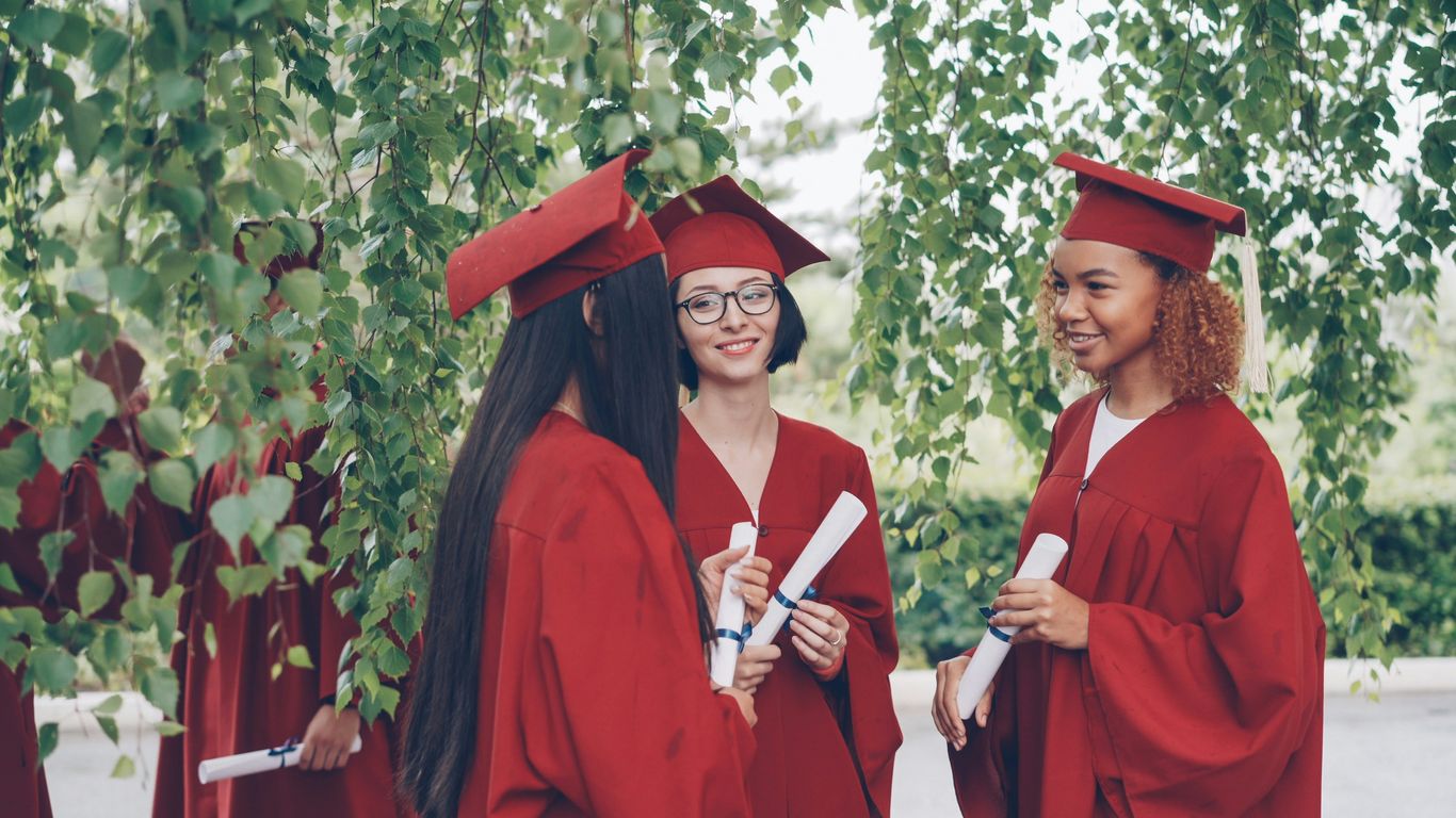 Graduates in red gowns holding diplomas converse outdoors.