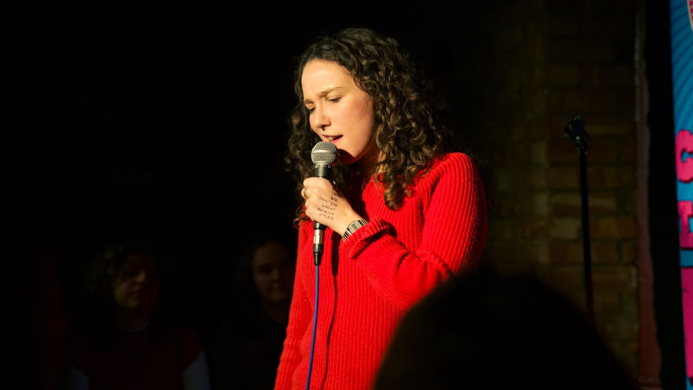 A person in a red sweater sings into a microphone on stage.