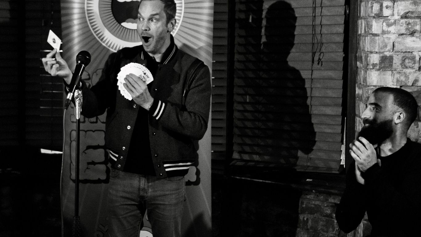 Magician performs a card trick with a surprised audience member watching.