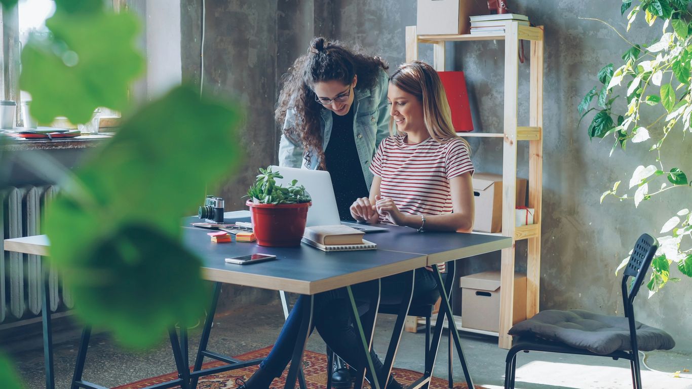 Two women collaborate at a laptop in an office.