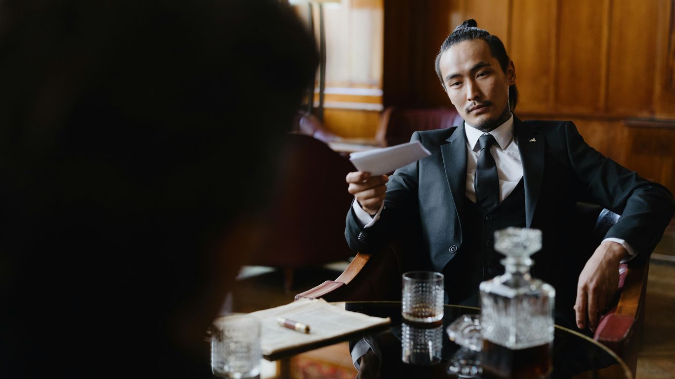 Man in suit offers paper, whiskey decanter and glass on table.