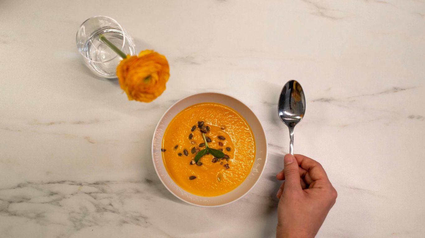 Bowl of orange soup, hand with spoon, yellow flower vase.