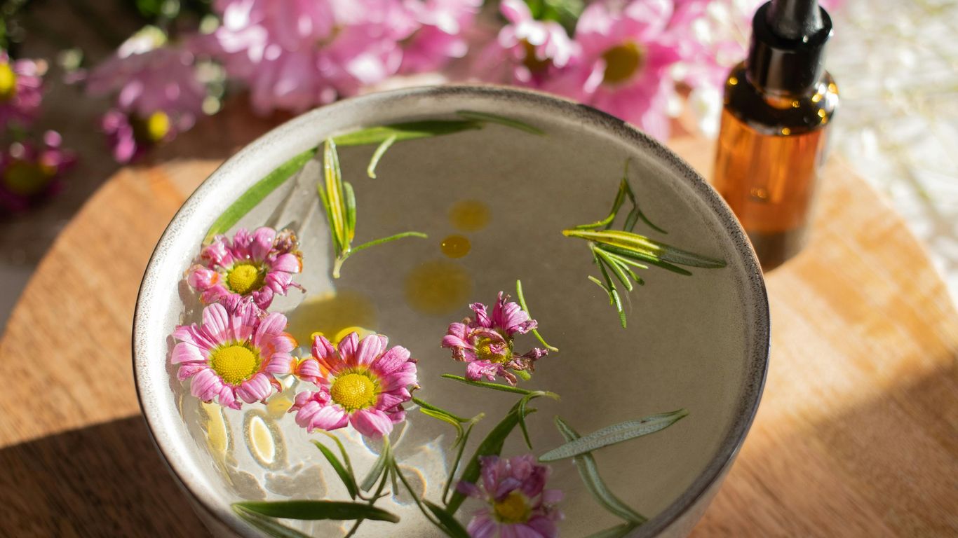 Bowl with floating flowers, essential oil bottle, wooden surface, sunlight.