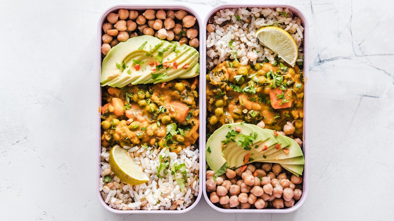 Two lunchboxes with rice, chickpeas, avocado, curry, lime slices.