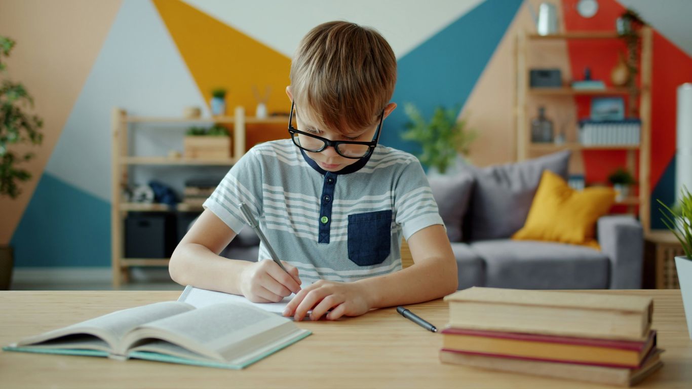 Young boy with glasses writing in a notebook at desk.