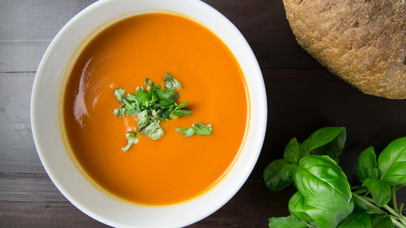 Bowl of tomato soup with herbs, bread, and basil leaves.