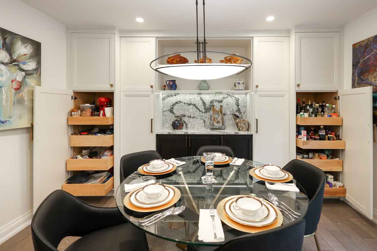 A modern dining room with a glass table, black chairs, and built-in pantry cabinets.