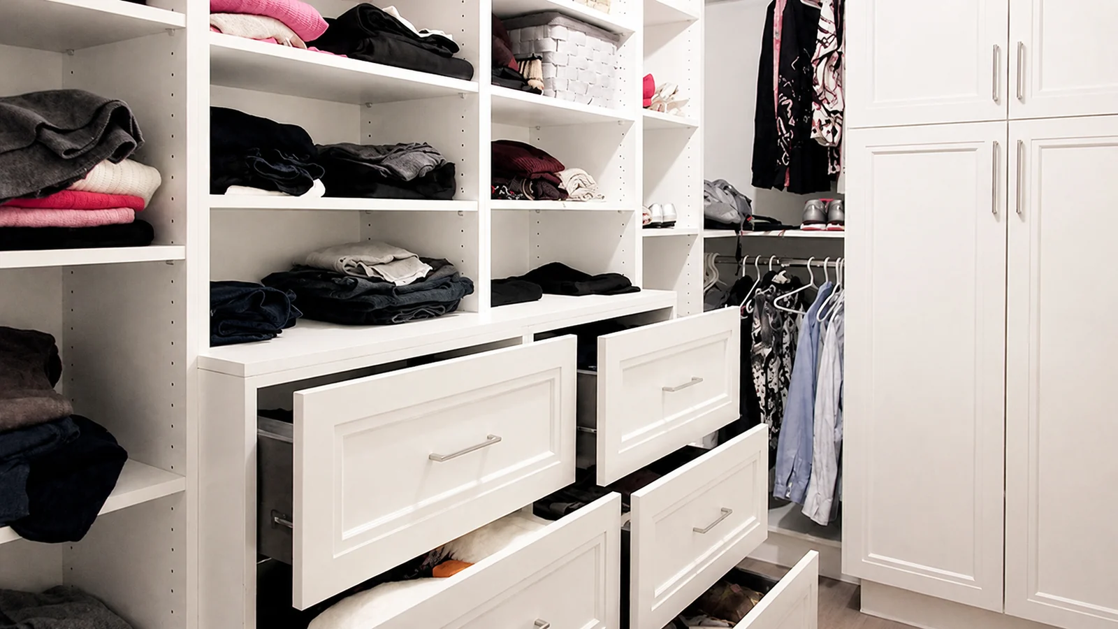 Neatly organized walk-in closet with white shelves, drawers, and hanging clothes.