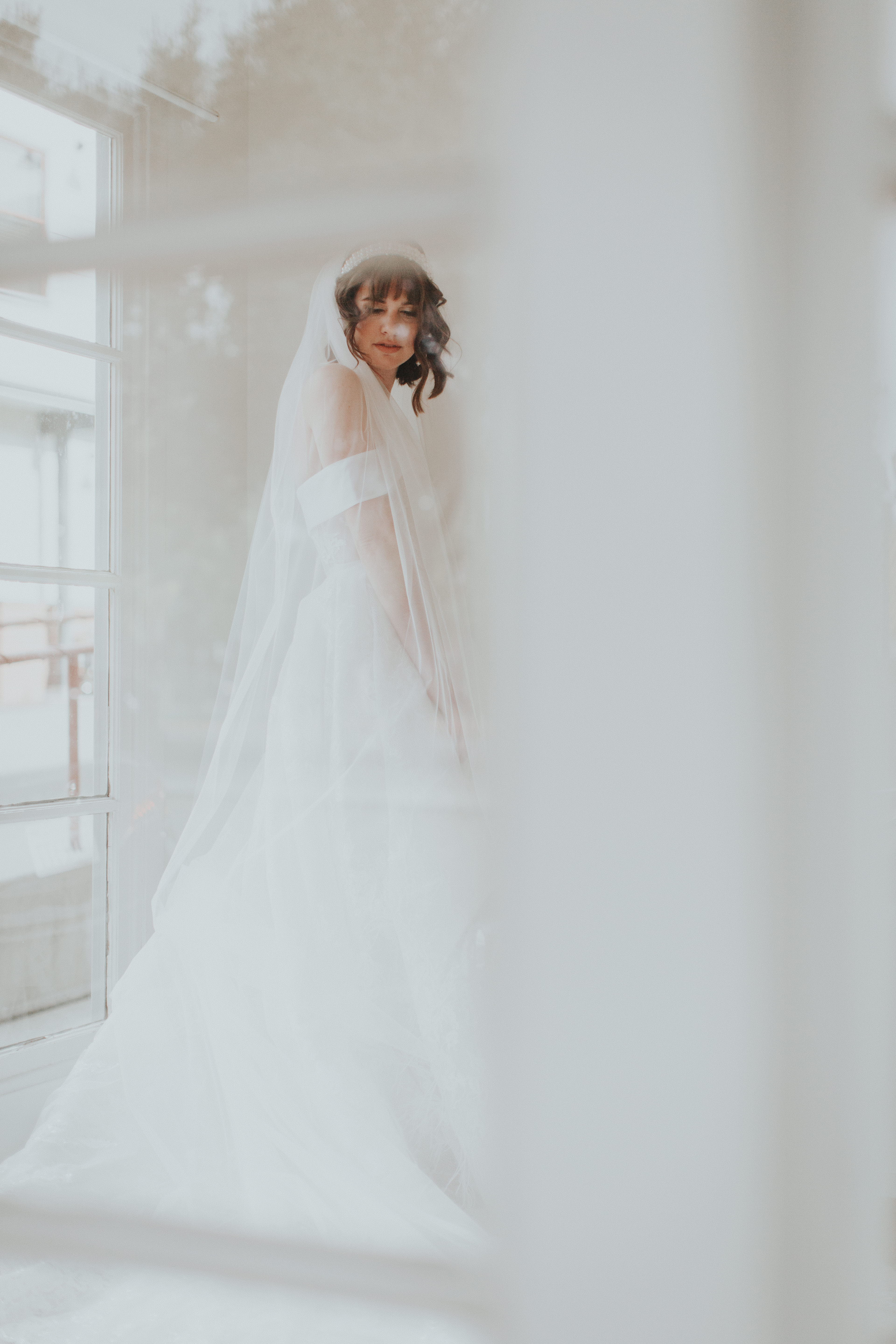 A person in a wedding dress stands near a window with a veil.