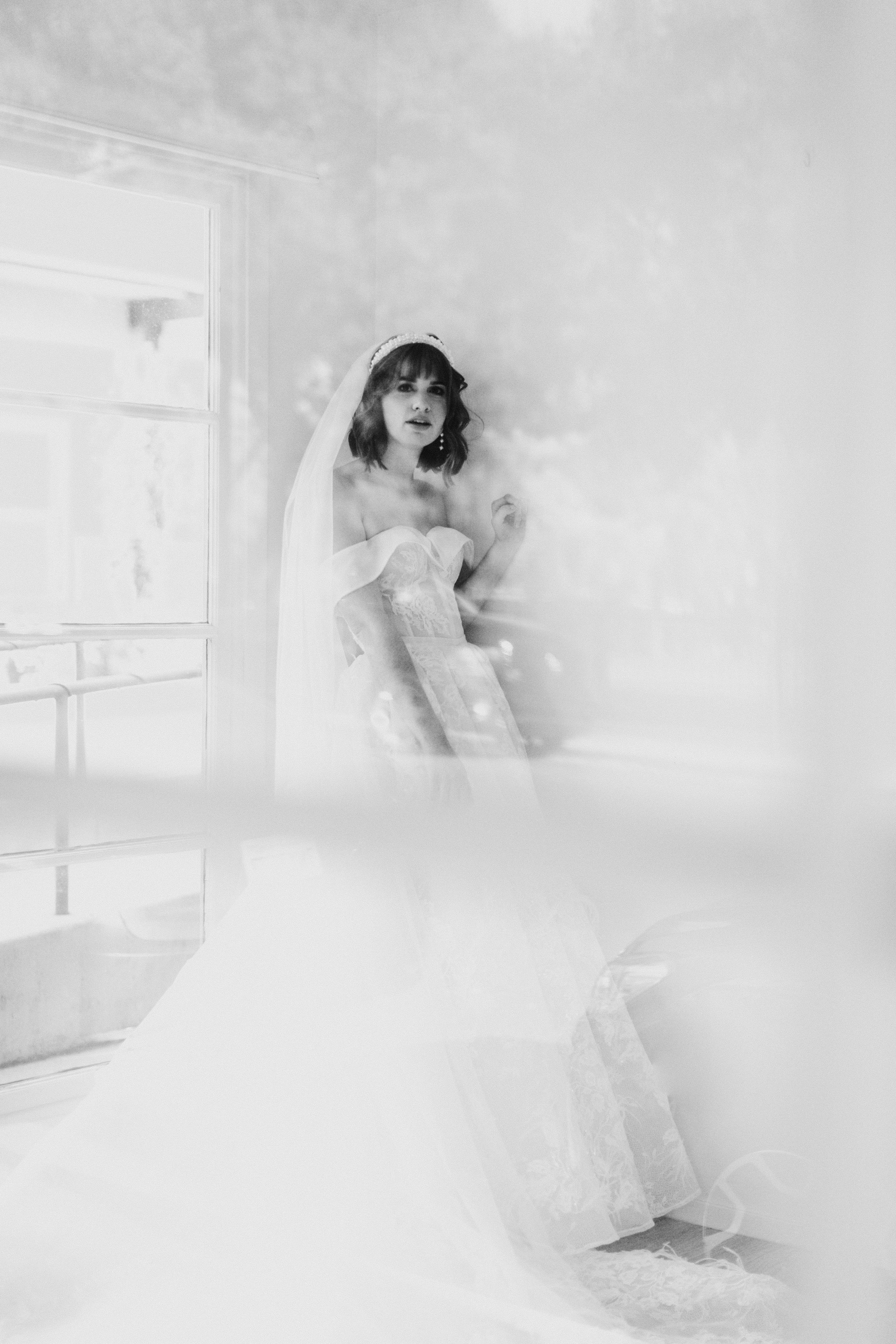 A person in a white wedding dress stands near a window with a veil.