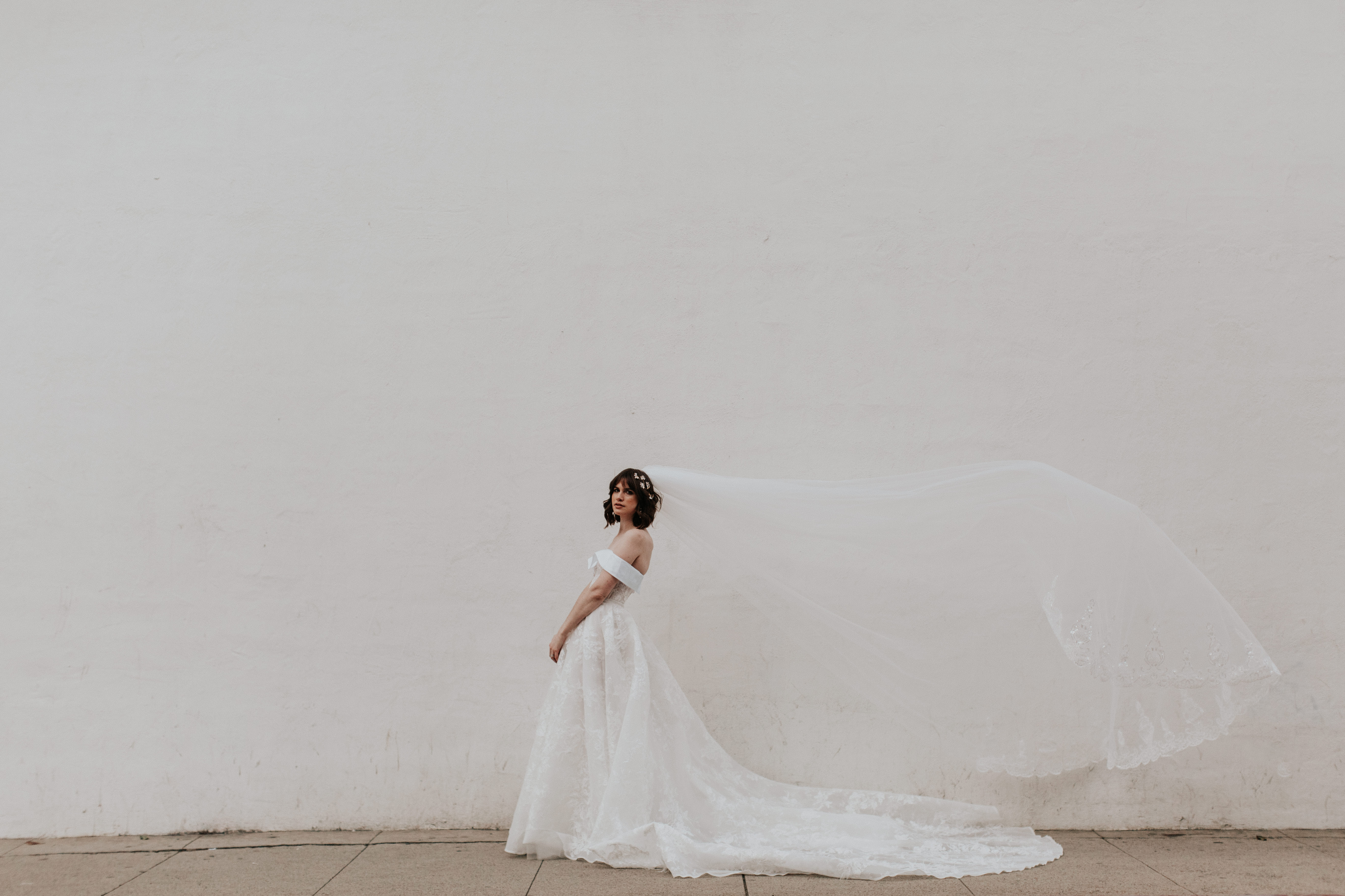 A person in a wedding dress stands against a plain wall with a flowing veil