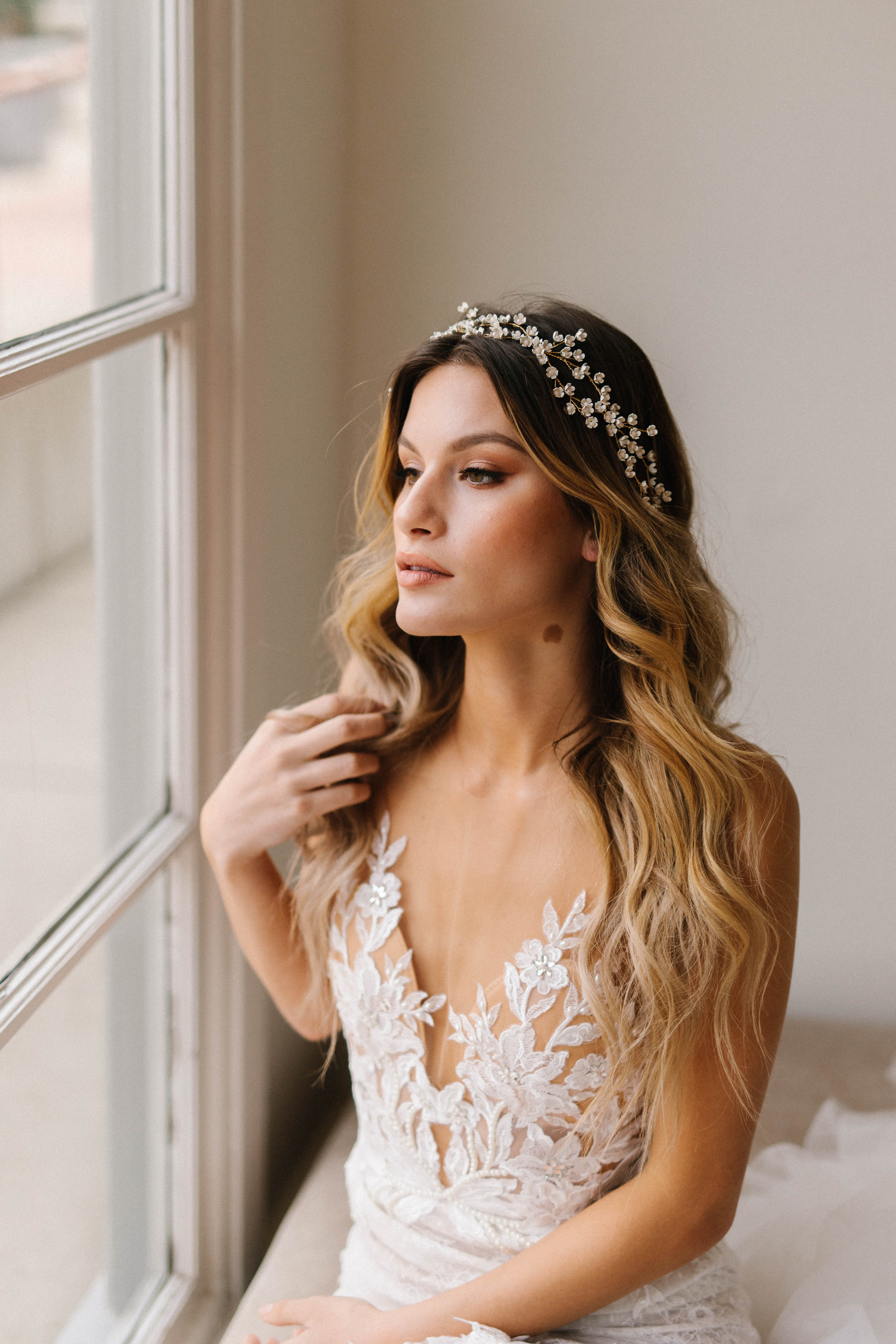 A bride in a white dress with a floral headpiece looks out a window.