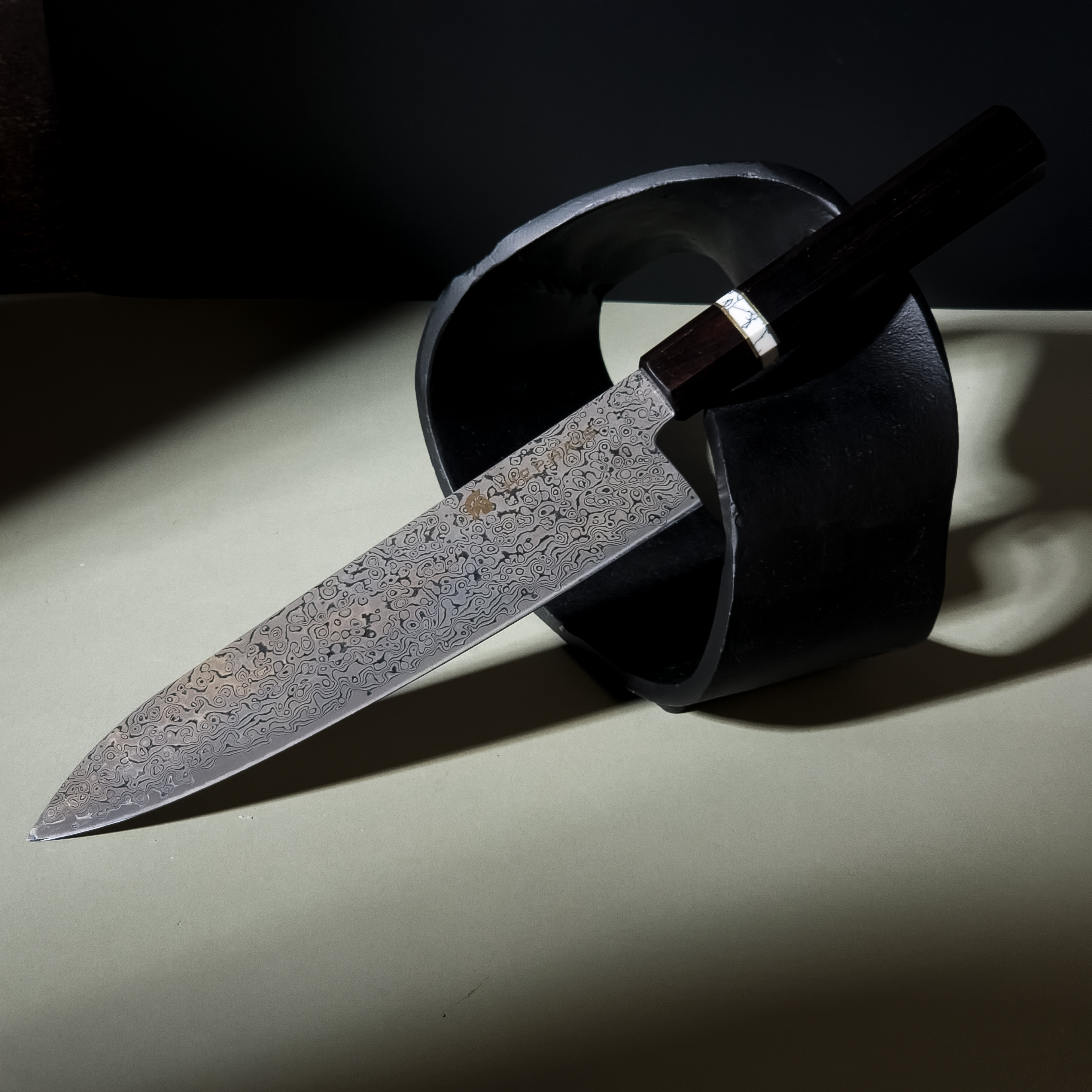 A close-up of a HEPHAIS Damascus Gyuto knife on a dark slate surface, highlighting the intricate blade pattern.
