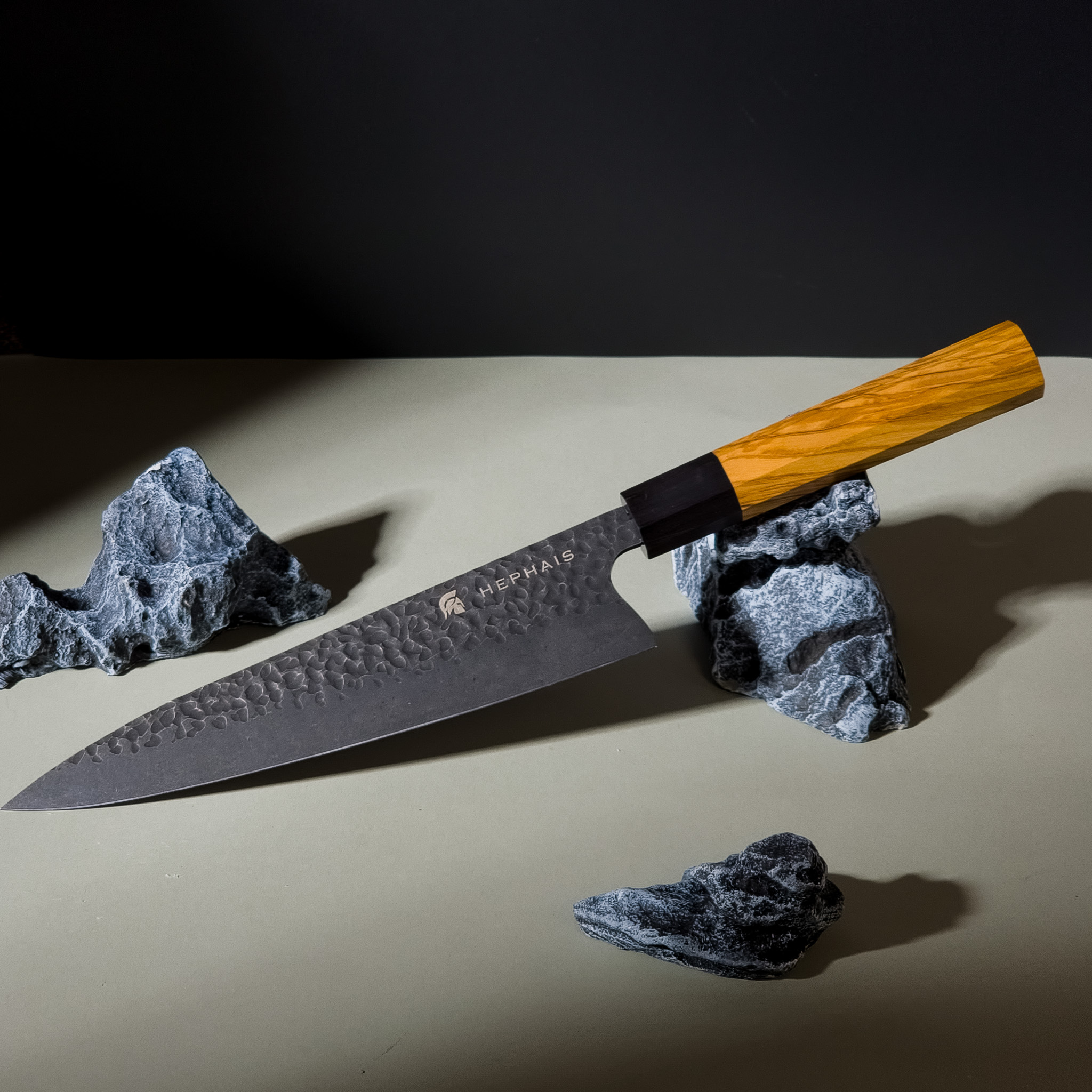 A hammered steel Hephais Perseus chef's knife with a wooden handle rests on rocks.