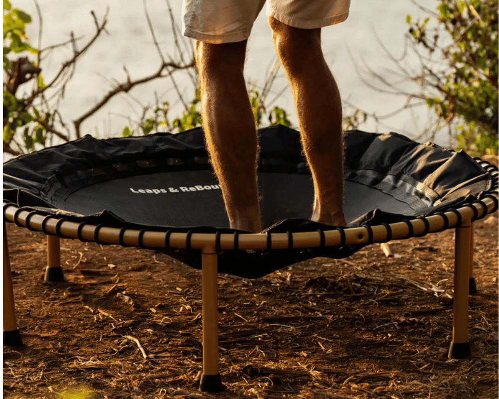 Person standing on a trampoline outdoors near water and trees.