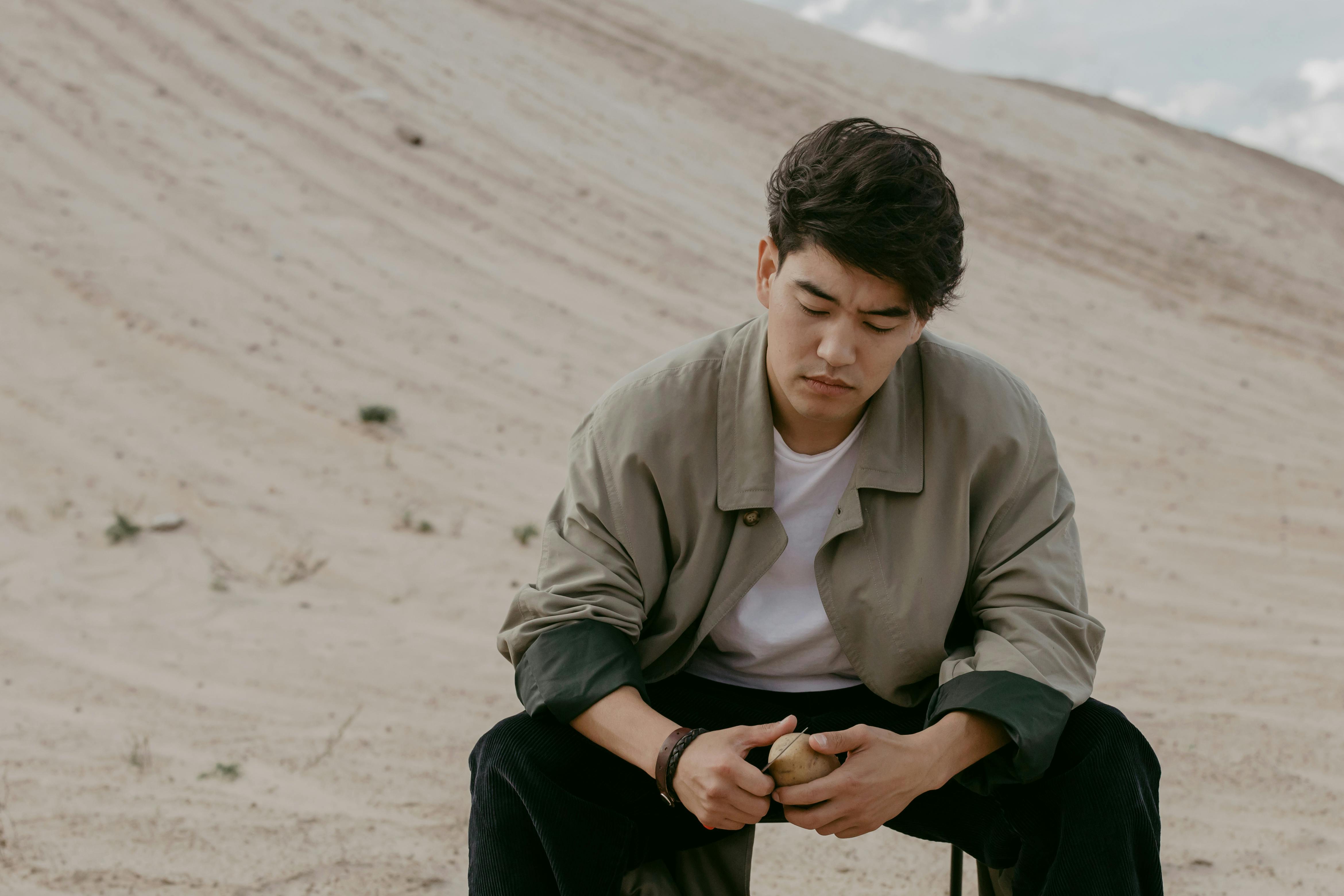 Person sitting on sand, peeling a potato, looking thoughtful.