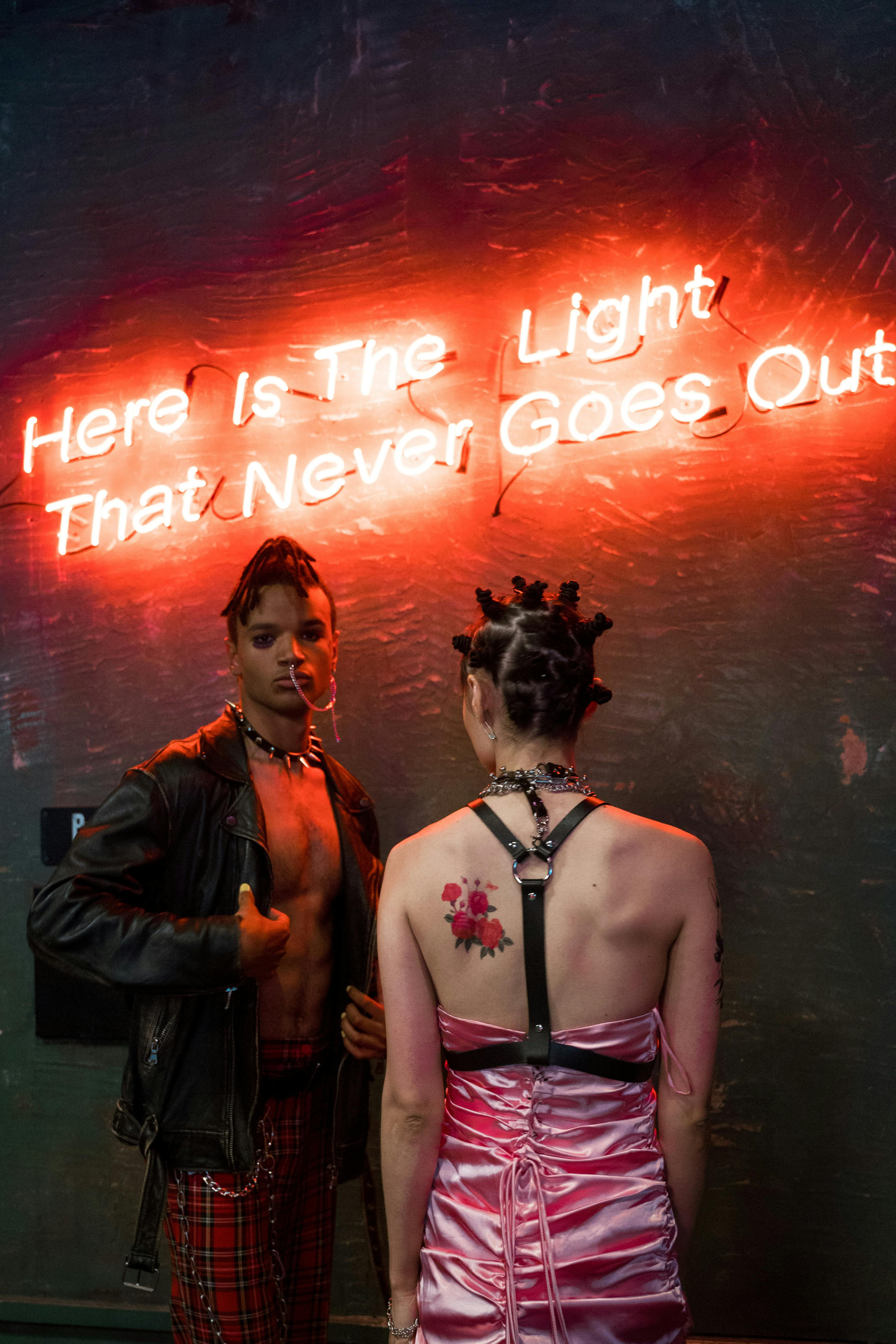 Two people in edgy outfits under neon sign, moody lighting.