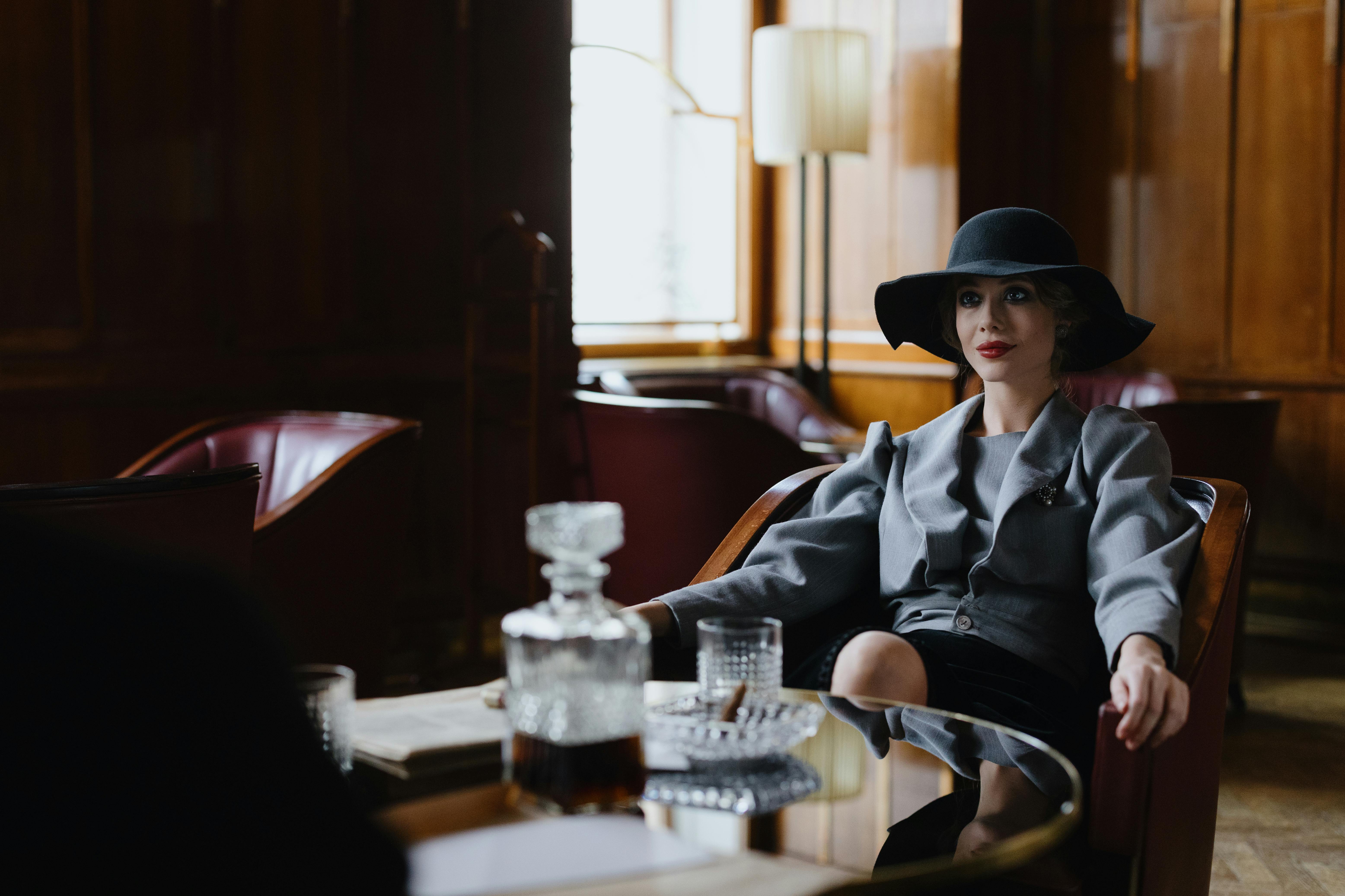 A person in a hat sits in a dimly lit room with a decanter.
