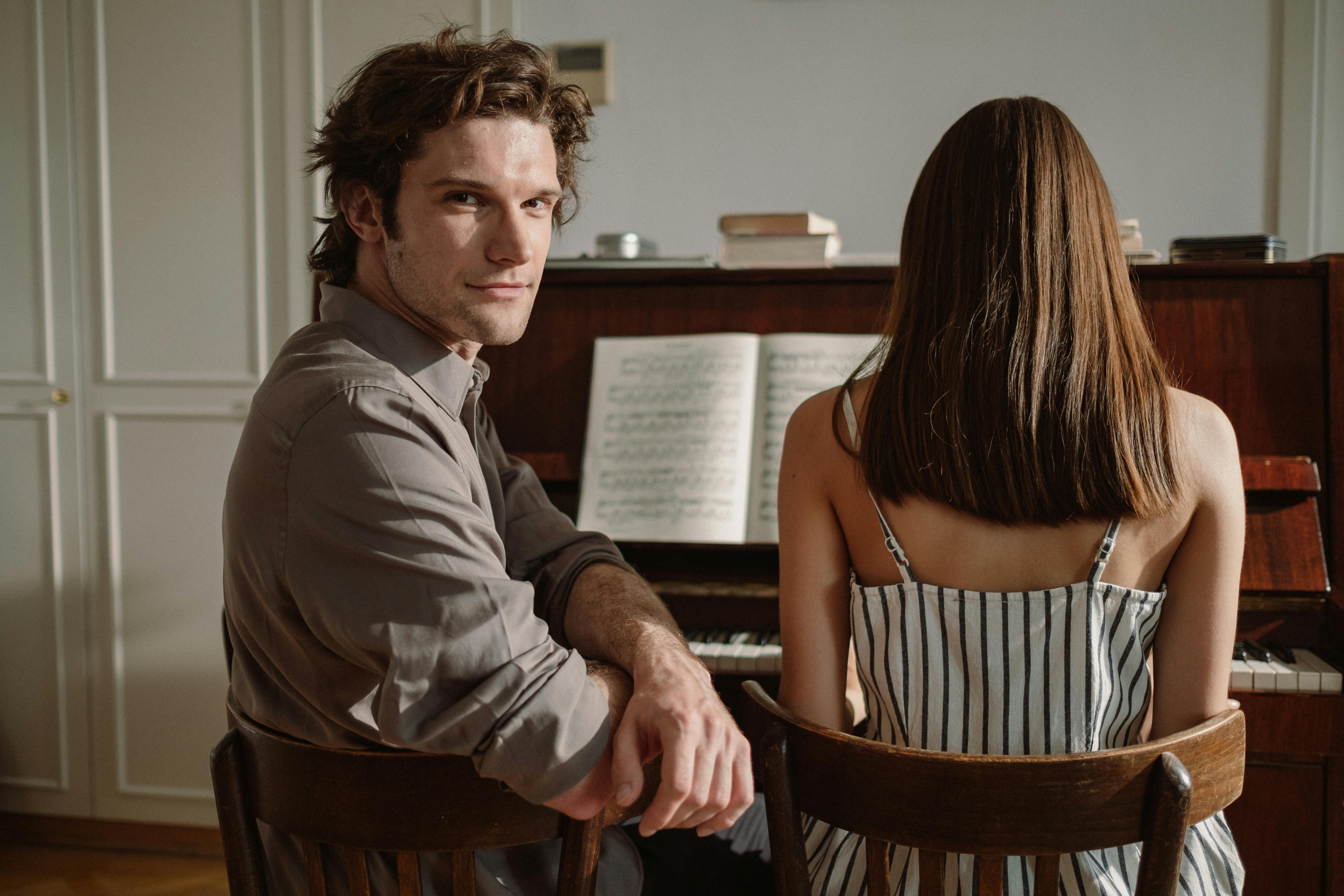 Two people sit at a piano with sheet music open.