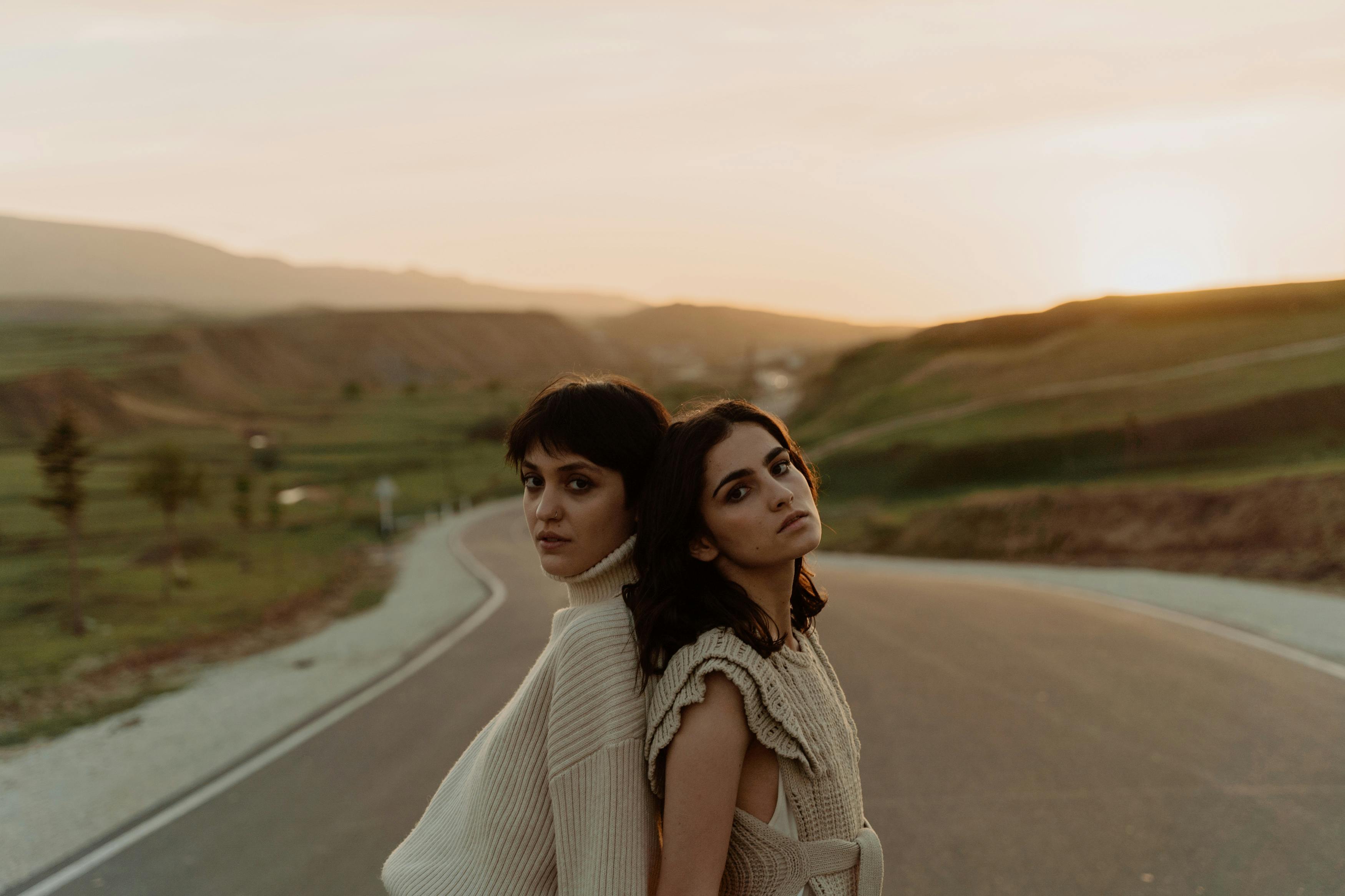 Two people stand back to back on a road at sunset.