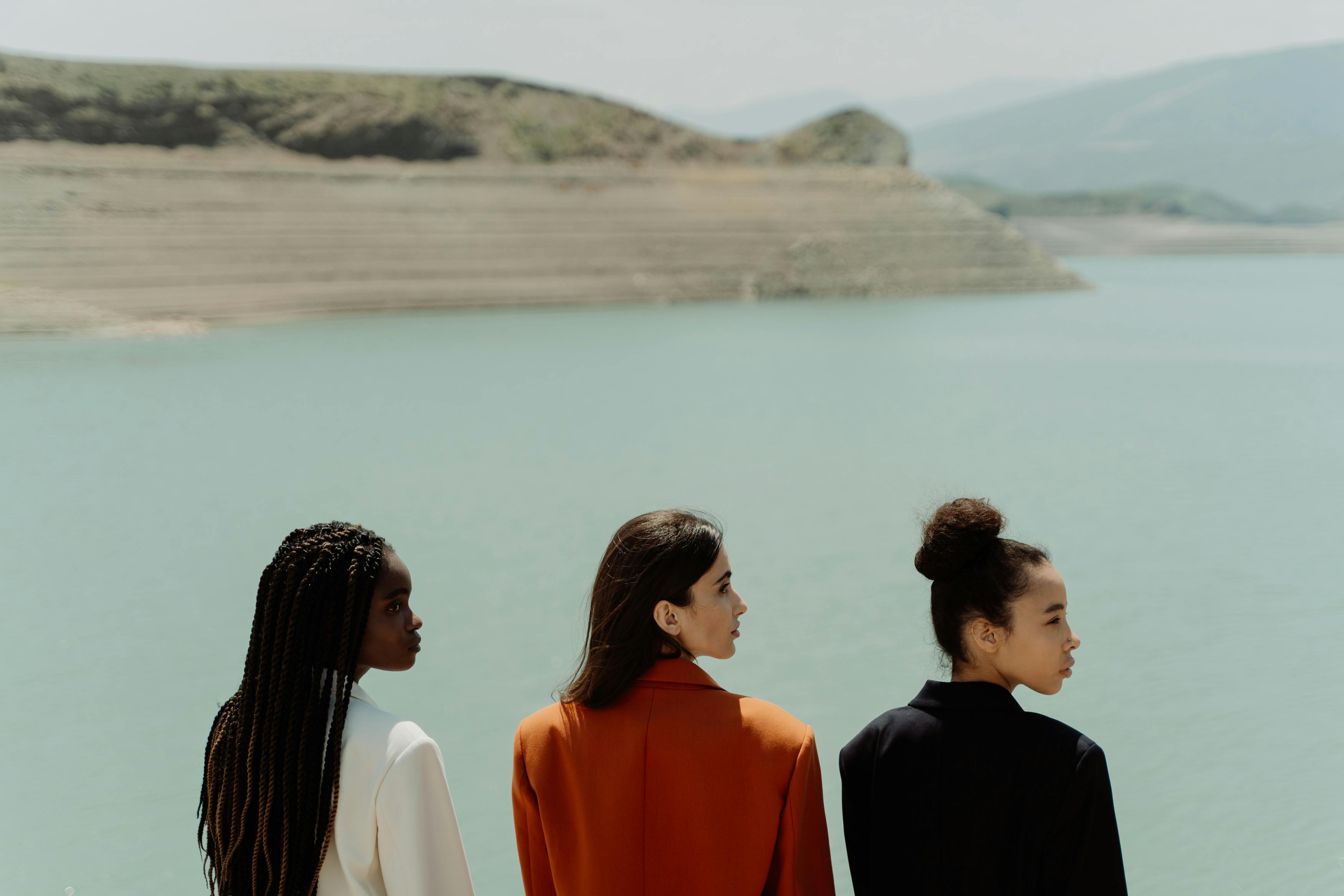 Three people stand facing a calm body of water and distant hills.