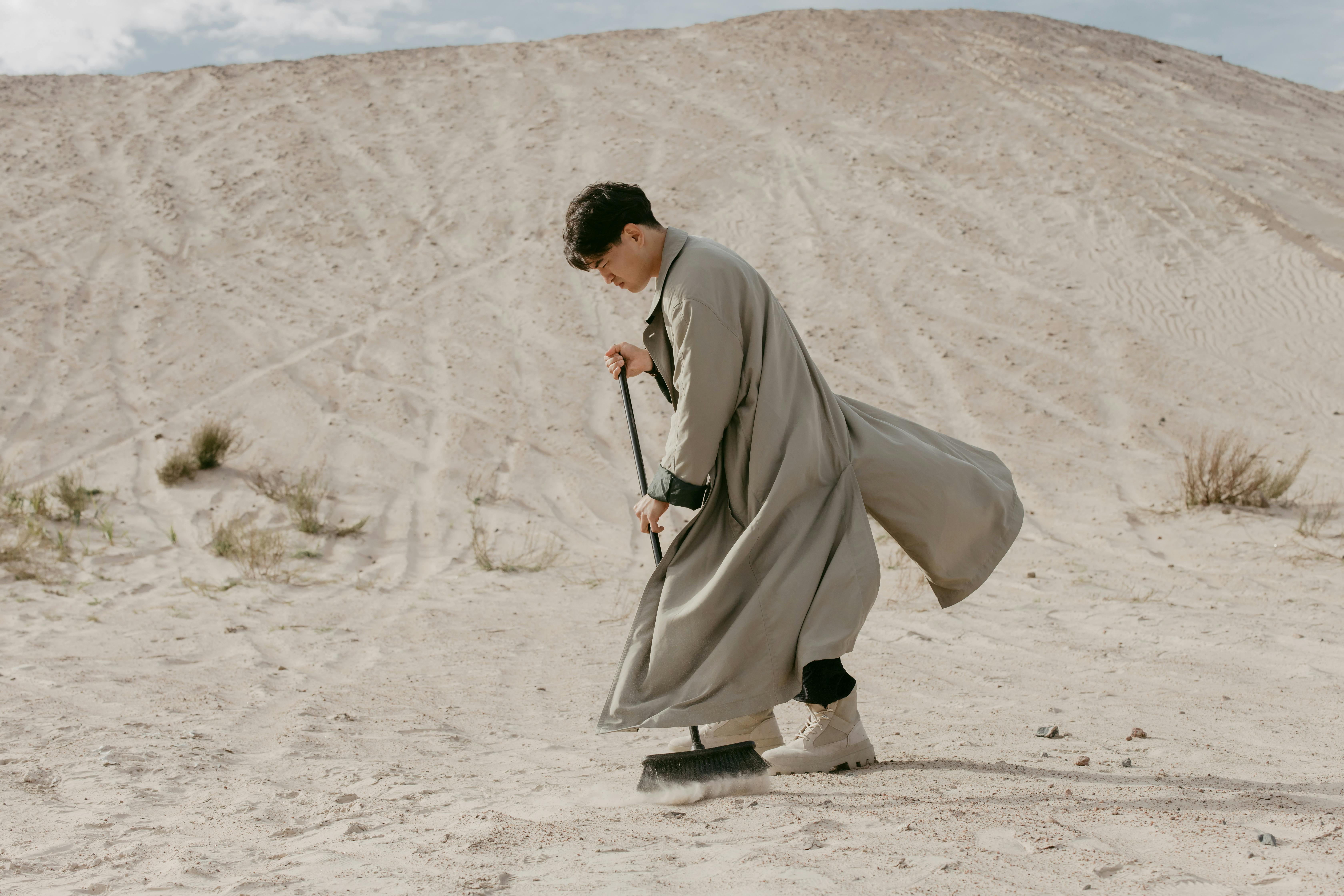 Person in long coat sweeping sand in a desert landscape.