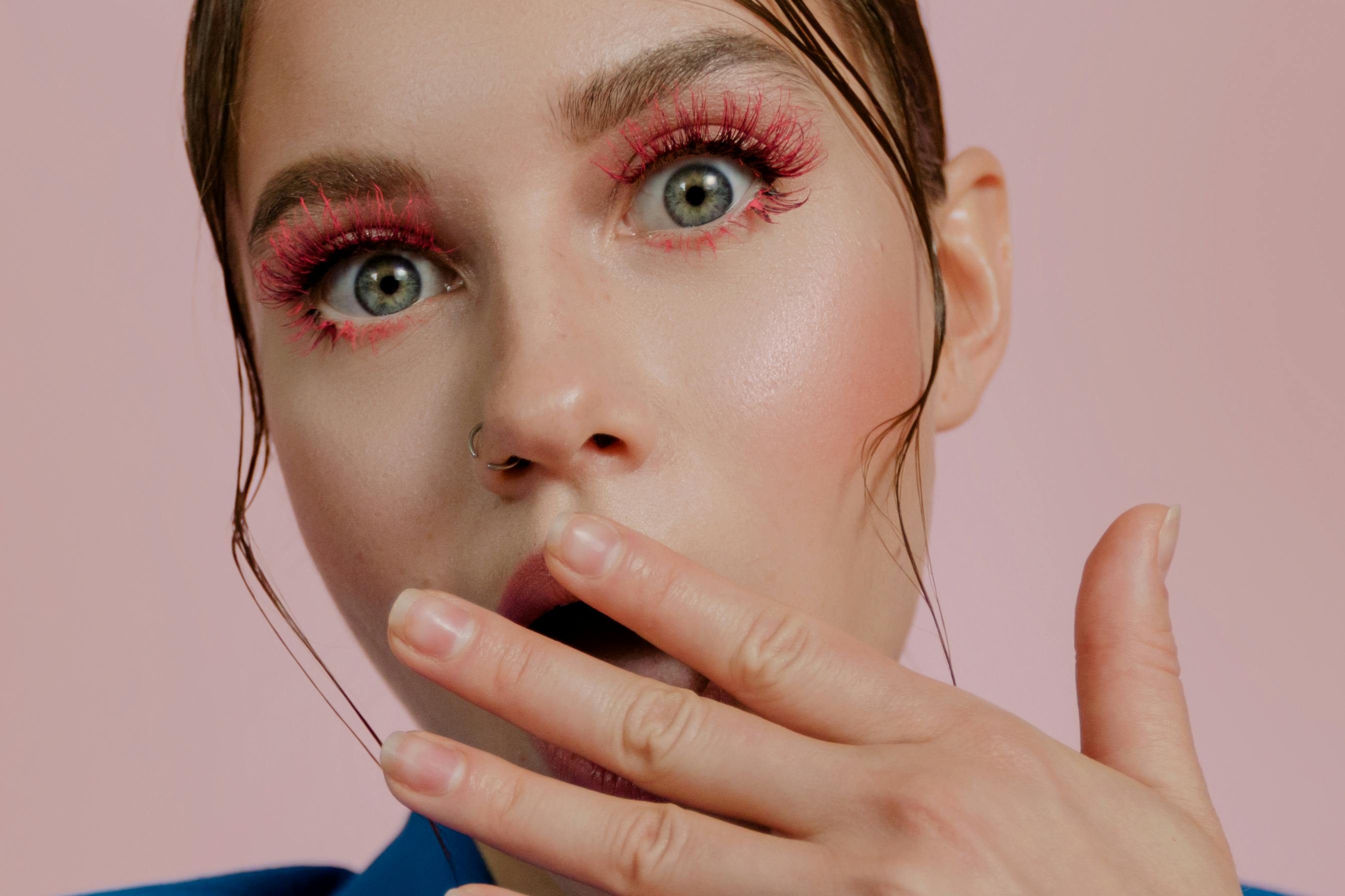 Person with surprised expression, pink makeup, hand near mouth.