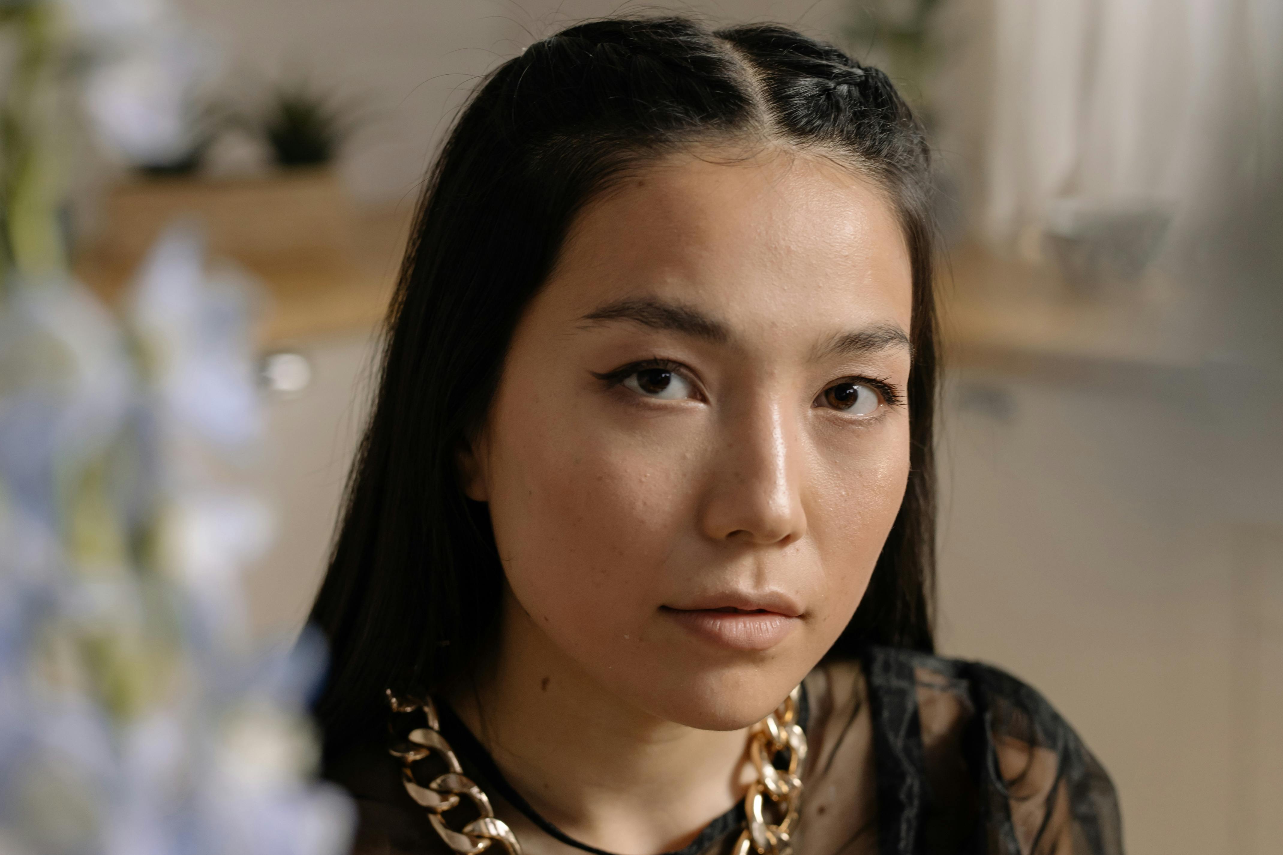 Person with long dark hair and gold chain necklace indoors.