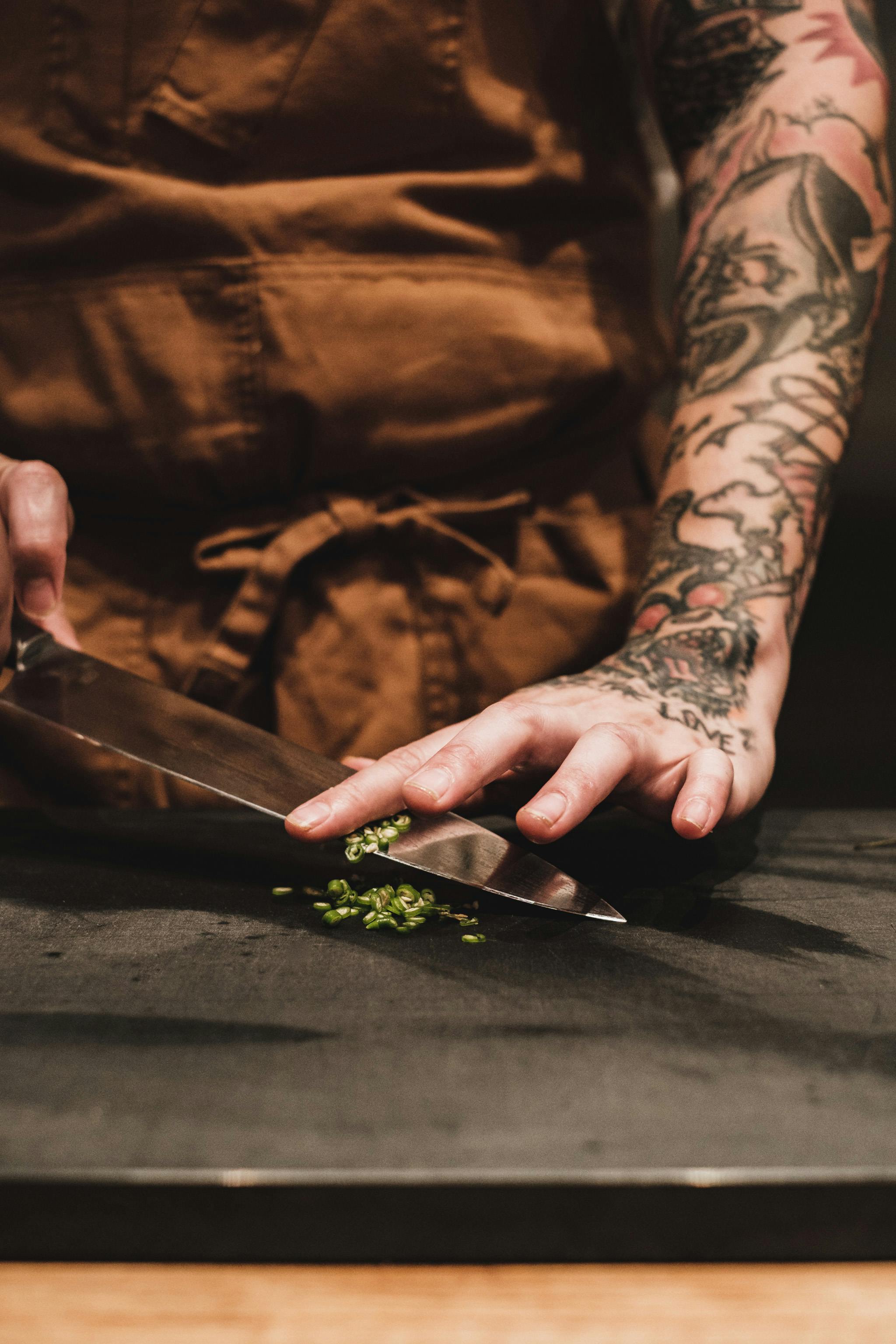 Tattooed person chopping green herbs with a large knife.