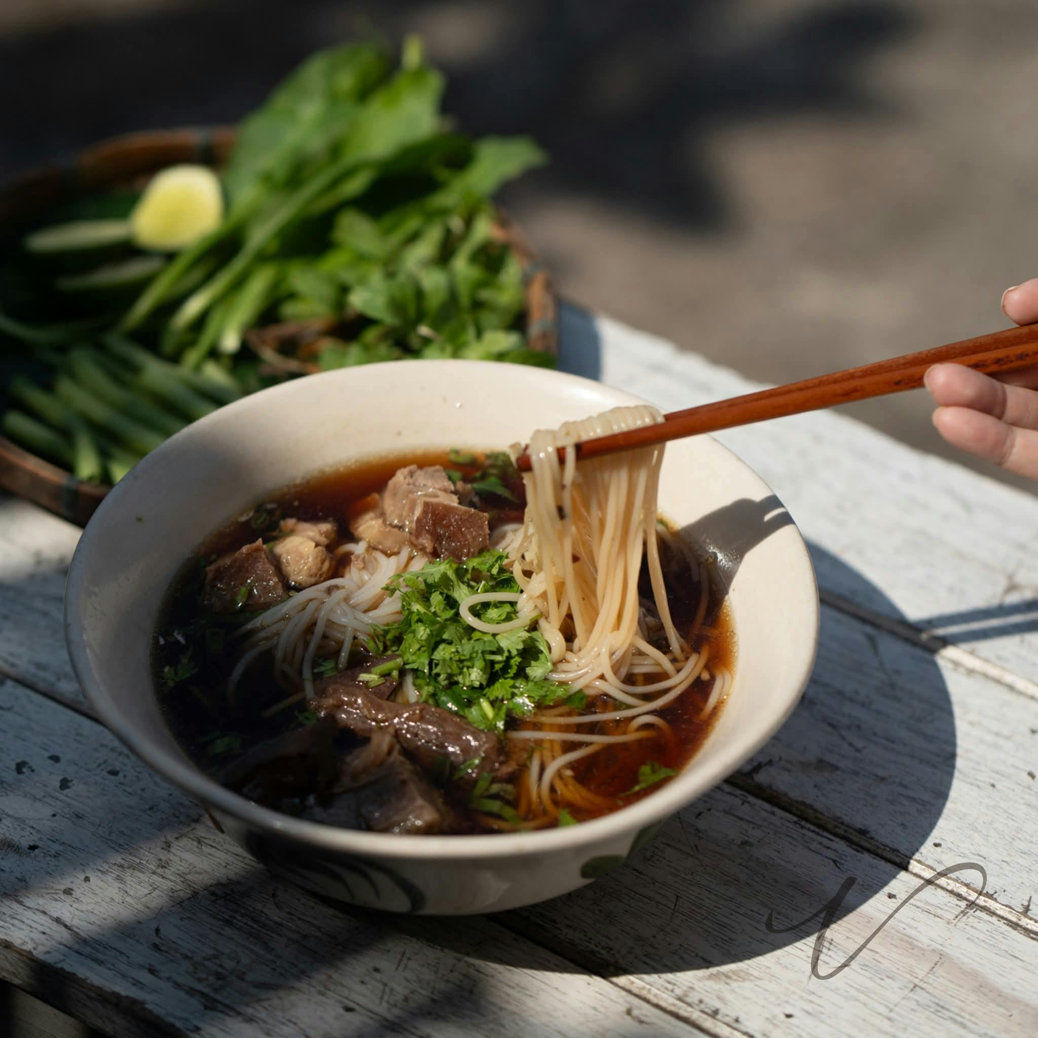 Bowl of noodle soup with herbs, chopsticks, and vegetables nearby.