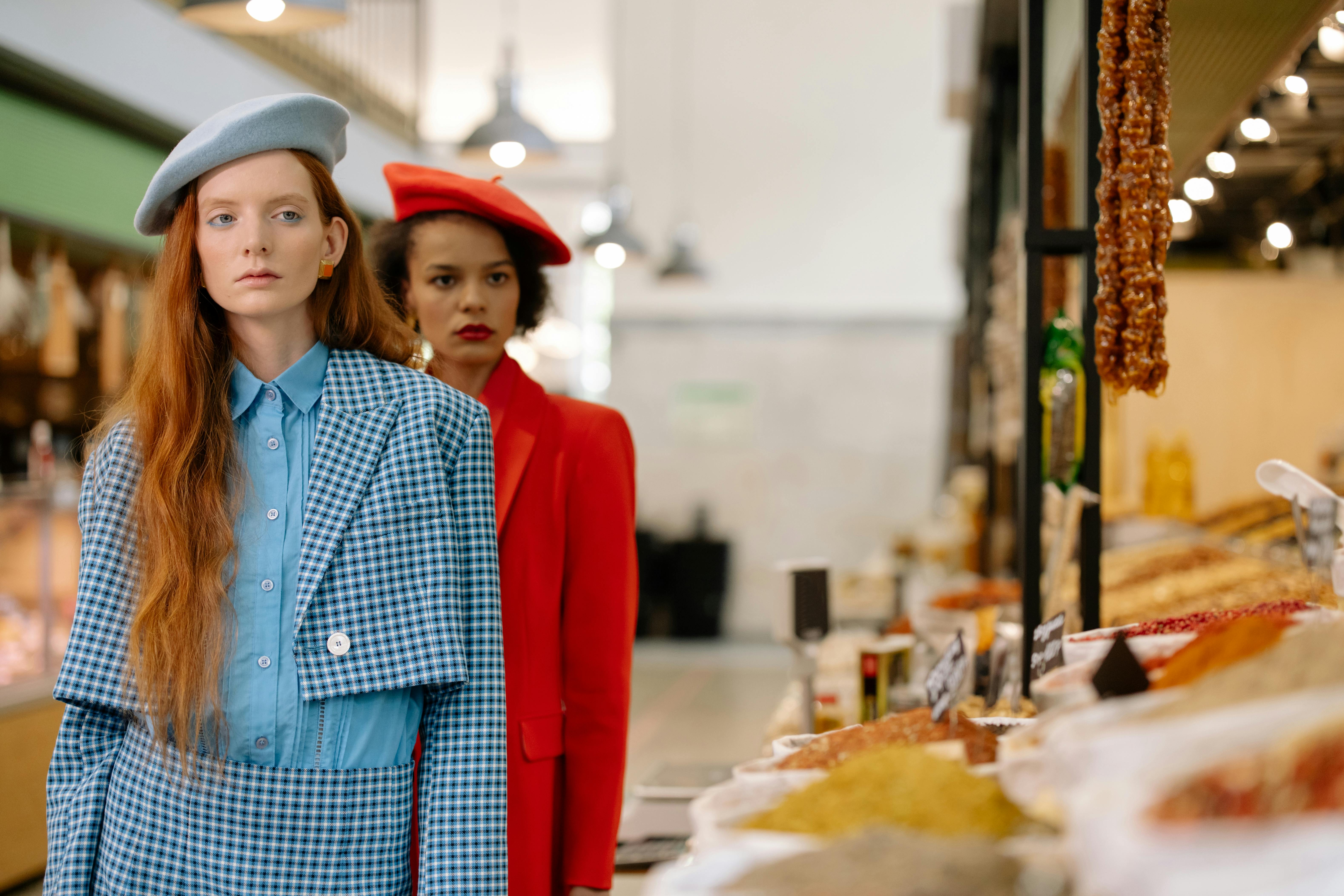 Two people in stylish outfits stand in a market with food.