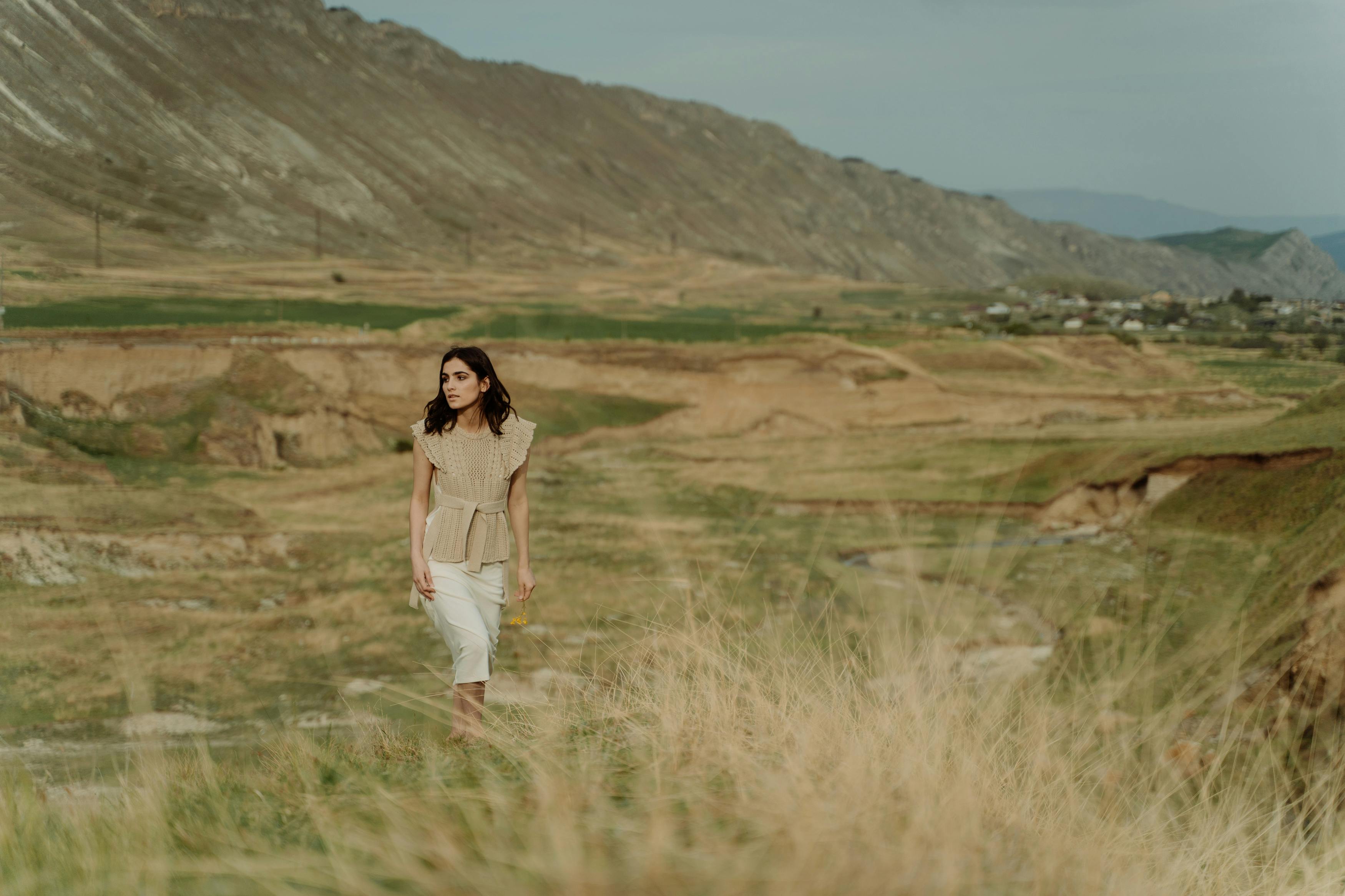 A person walks through a grassy field with mountains in the background.