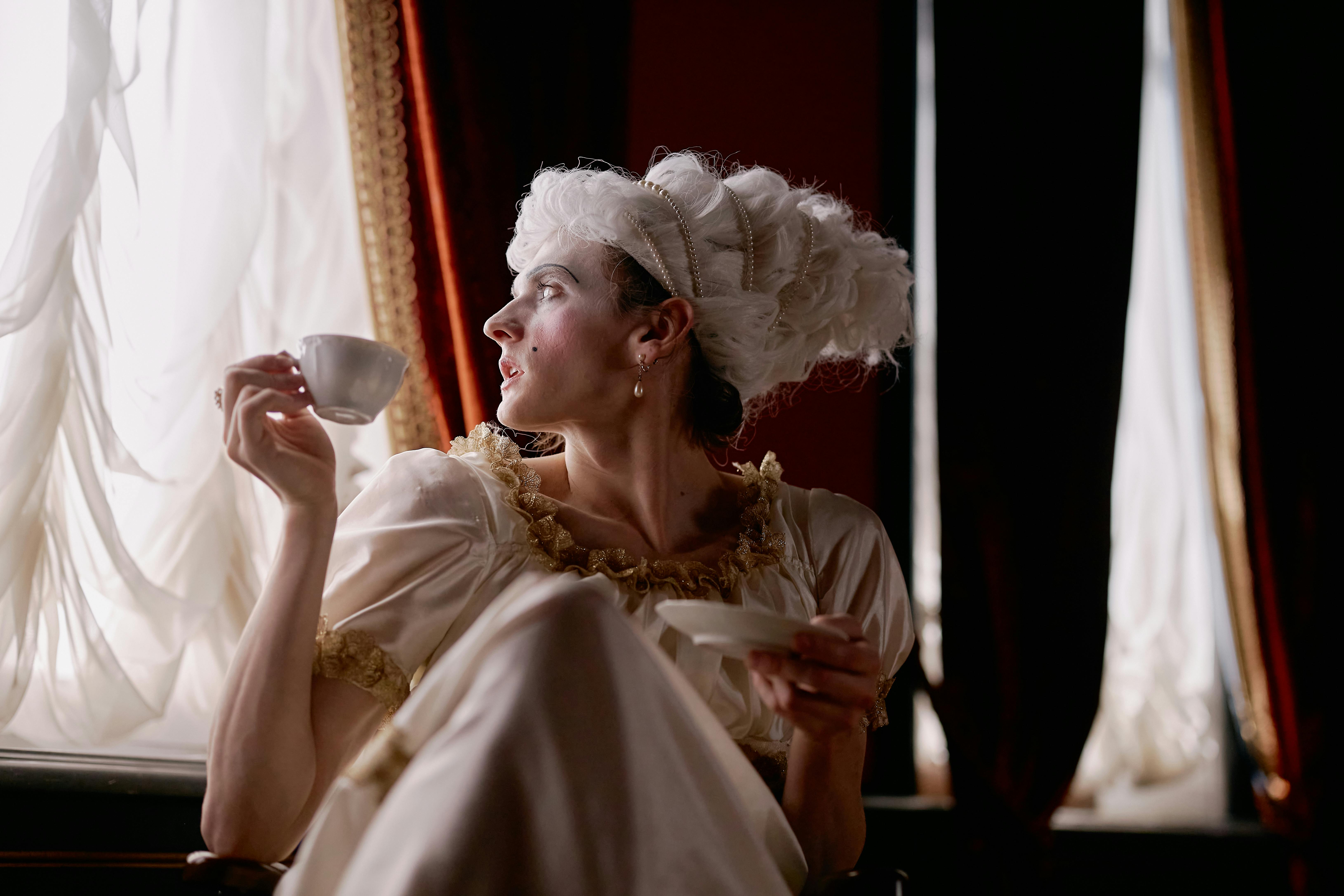 Person in historical costume drinking tea by elegant window curtains.