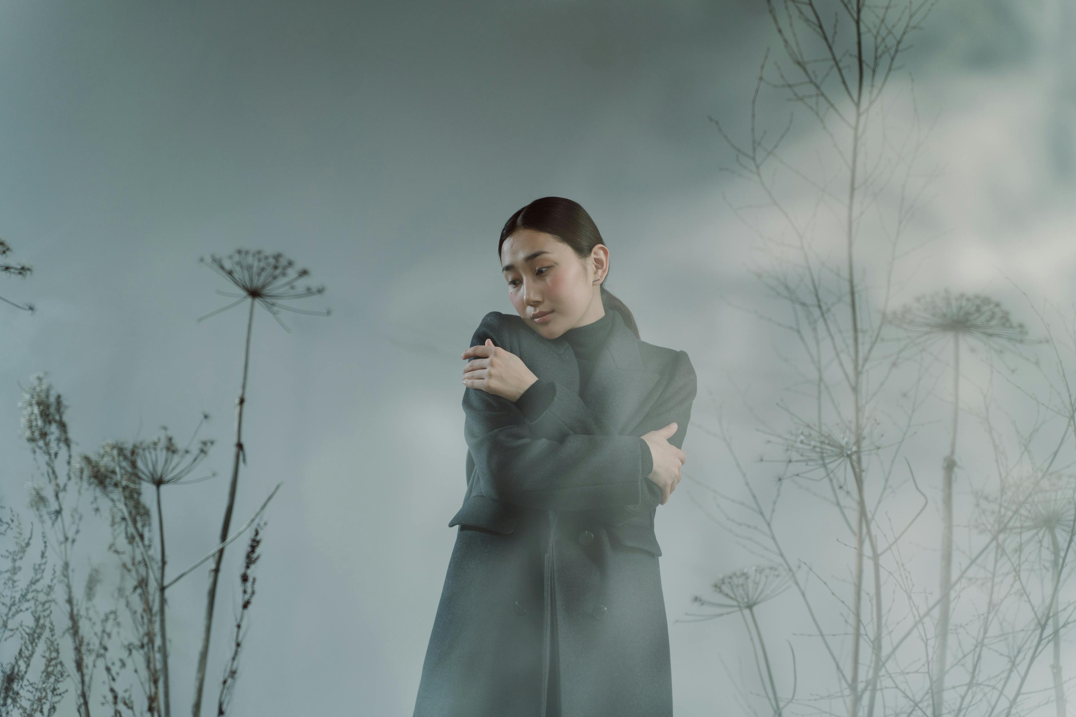 Person in dark coat standing among tall, dried plants, misty atmosphere.